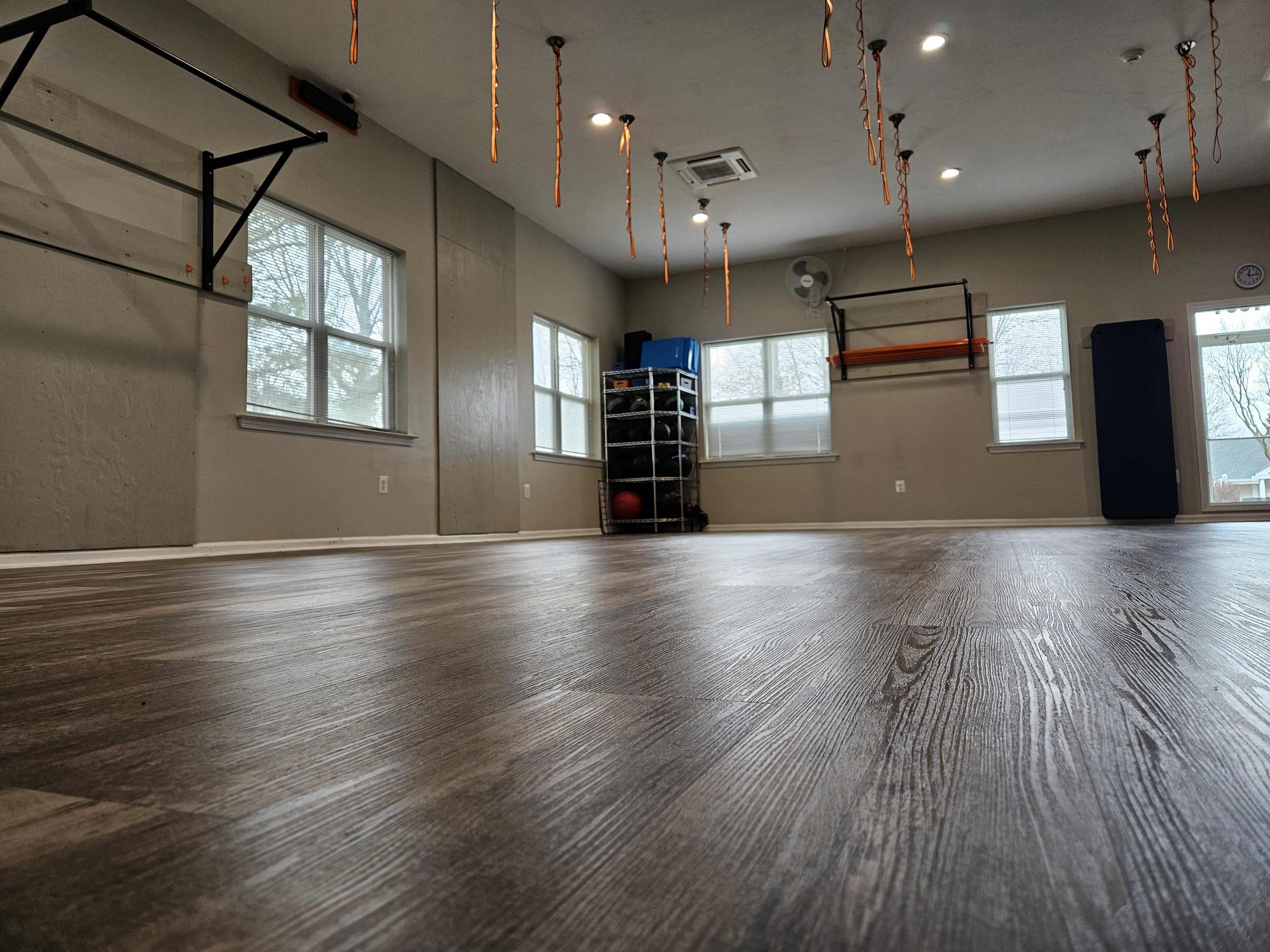 An empty gym space with wooden flooring, wall-mounted fitness equipment, and natural light coming through the windows.