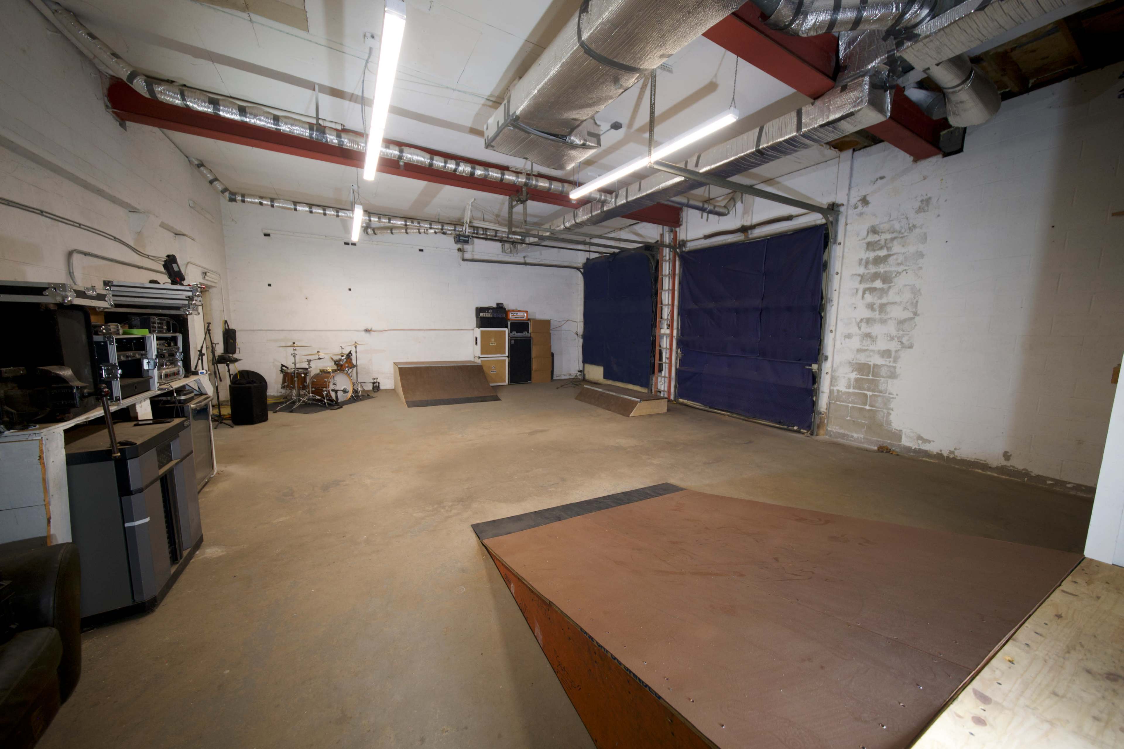 An empty indoor space with two ramps, a drum set, and exposed beams, illuminated by overhead fluorescent lights.