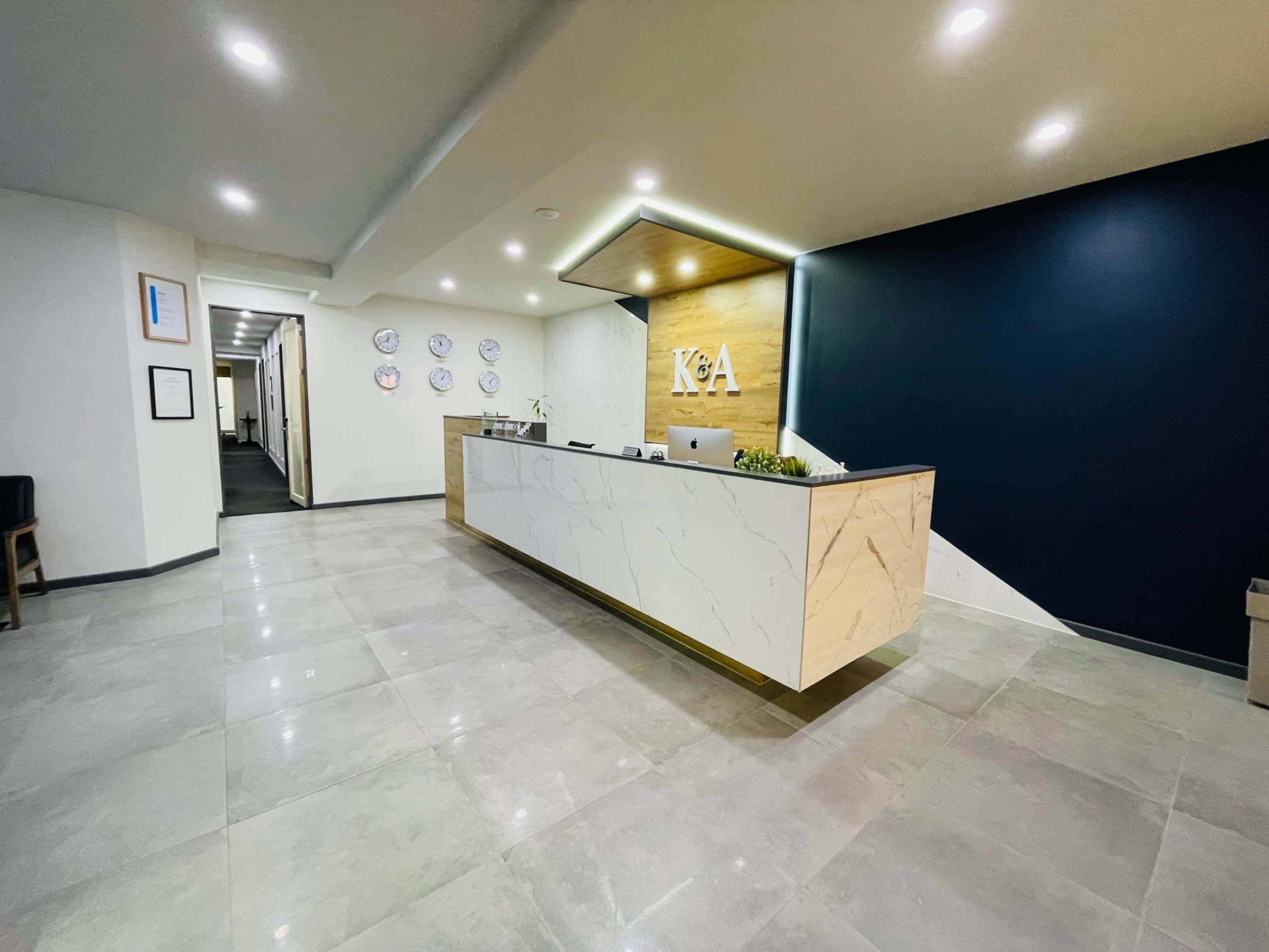 The image shows a modern reception area with a marble front desk featuring the letters "K&A" and a dark wall behind it, with a hallway leading to other rooms in the background.
