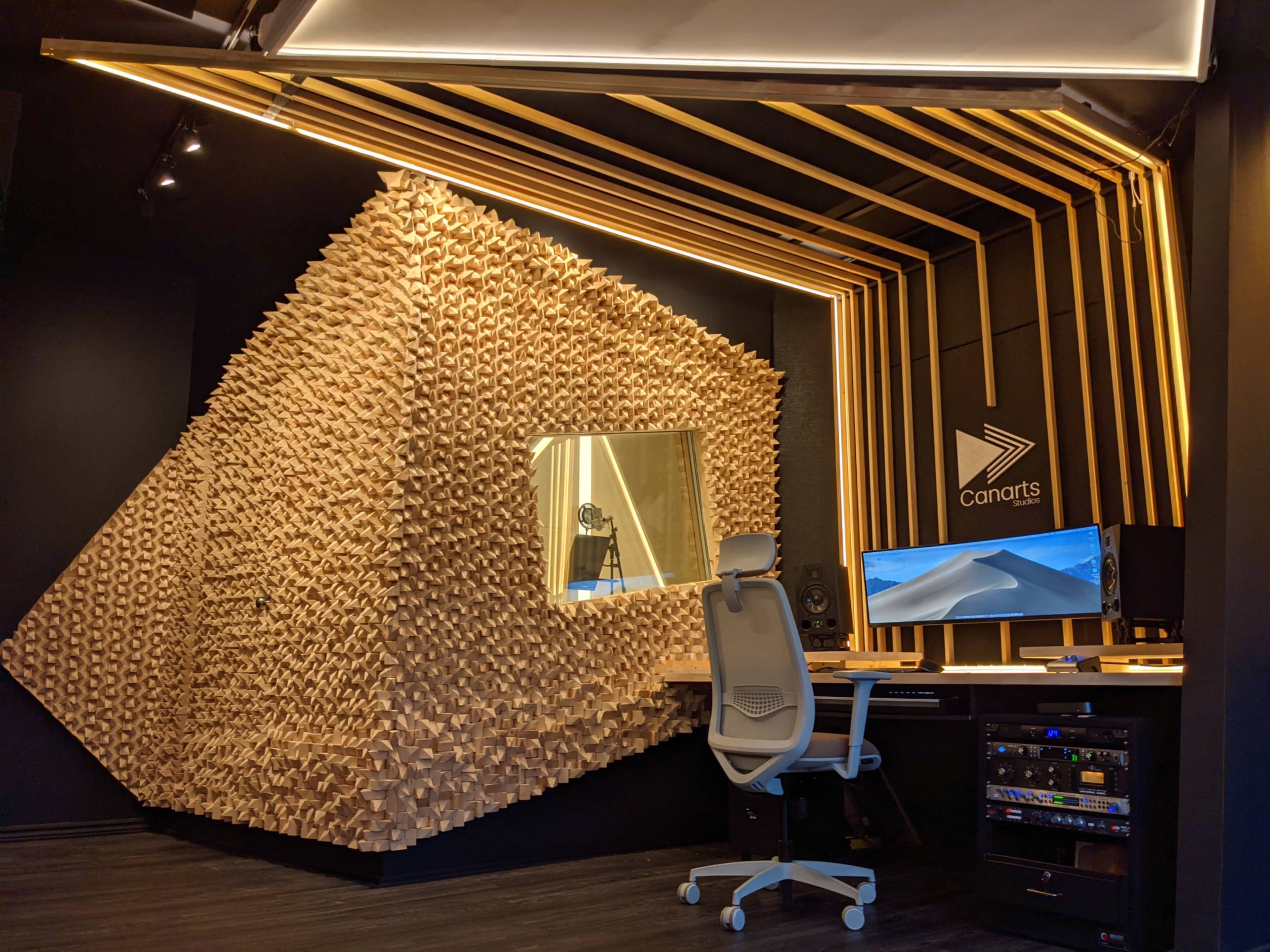 A modern audio studio featuring a unique wooden soundproof wall, a desk with a computer setup, and ambient lighting.