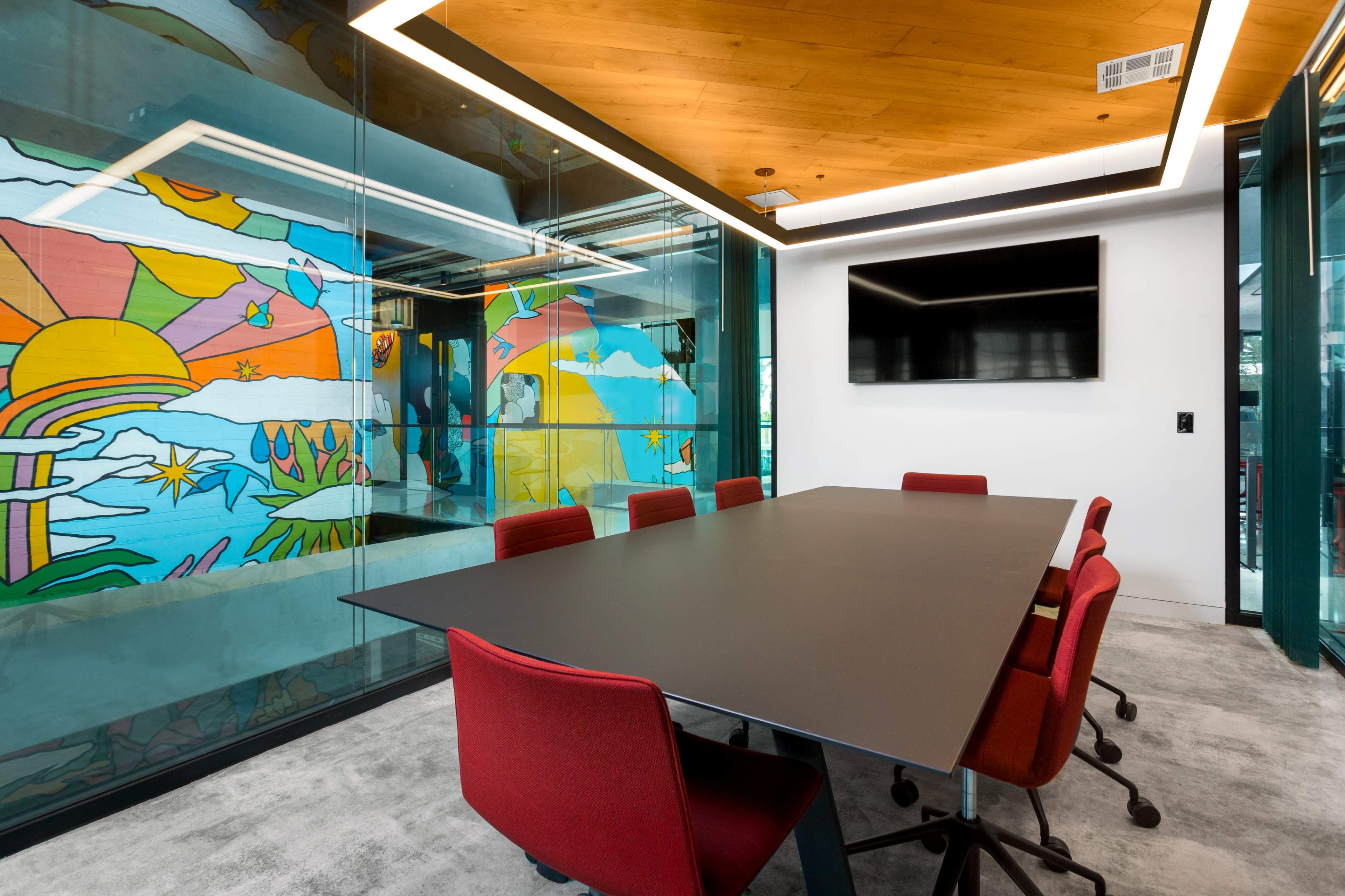 A modern conference room features a large black table surrounded by red chairs, with glass walls displaying a colorful mural and a mounted television.