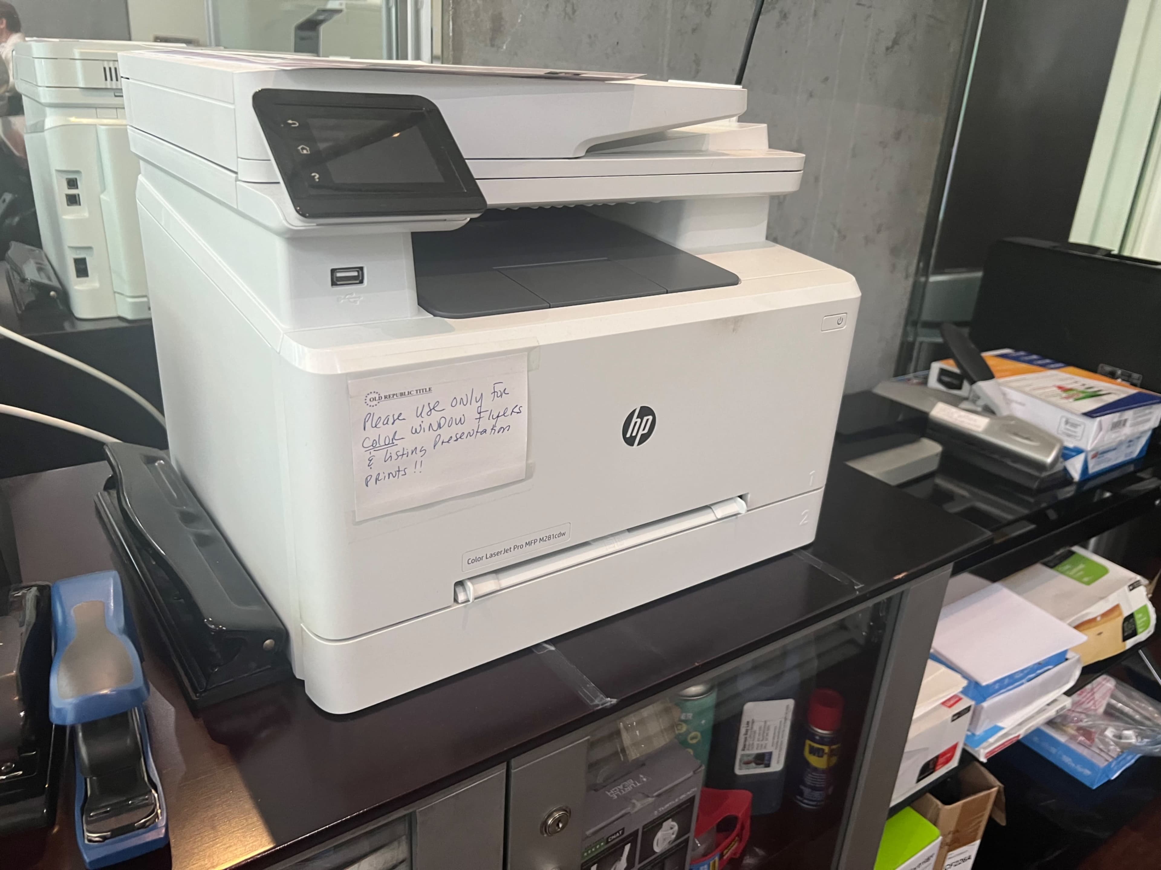 A white HP printer is placed on a black desk, surrounded by various office supplies and equipment.