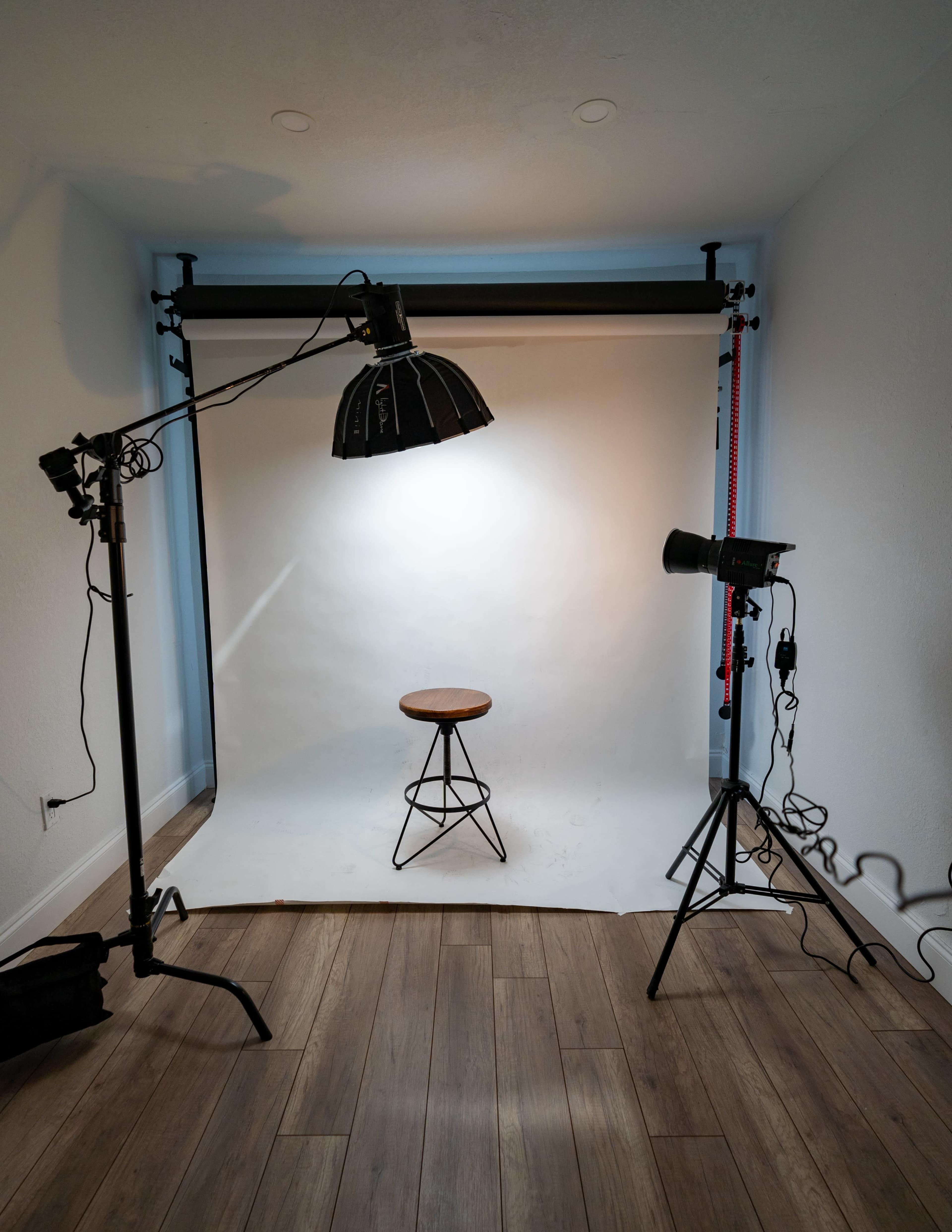 A minimalist photography studio setup features a wooden stool centered under a large overhead light, with two adjustable light stands on either side and a plain backdrop.