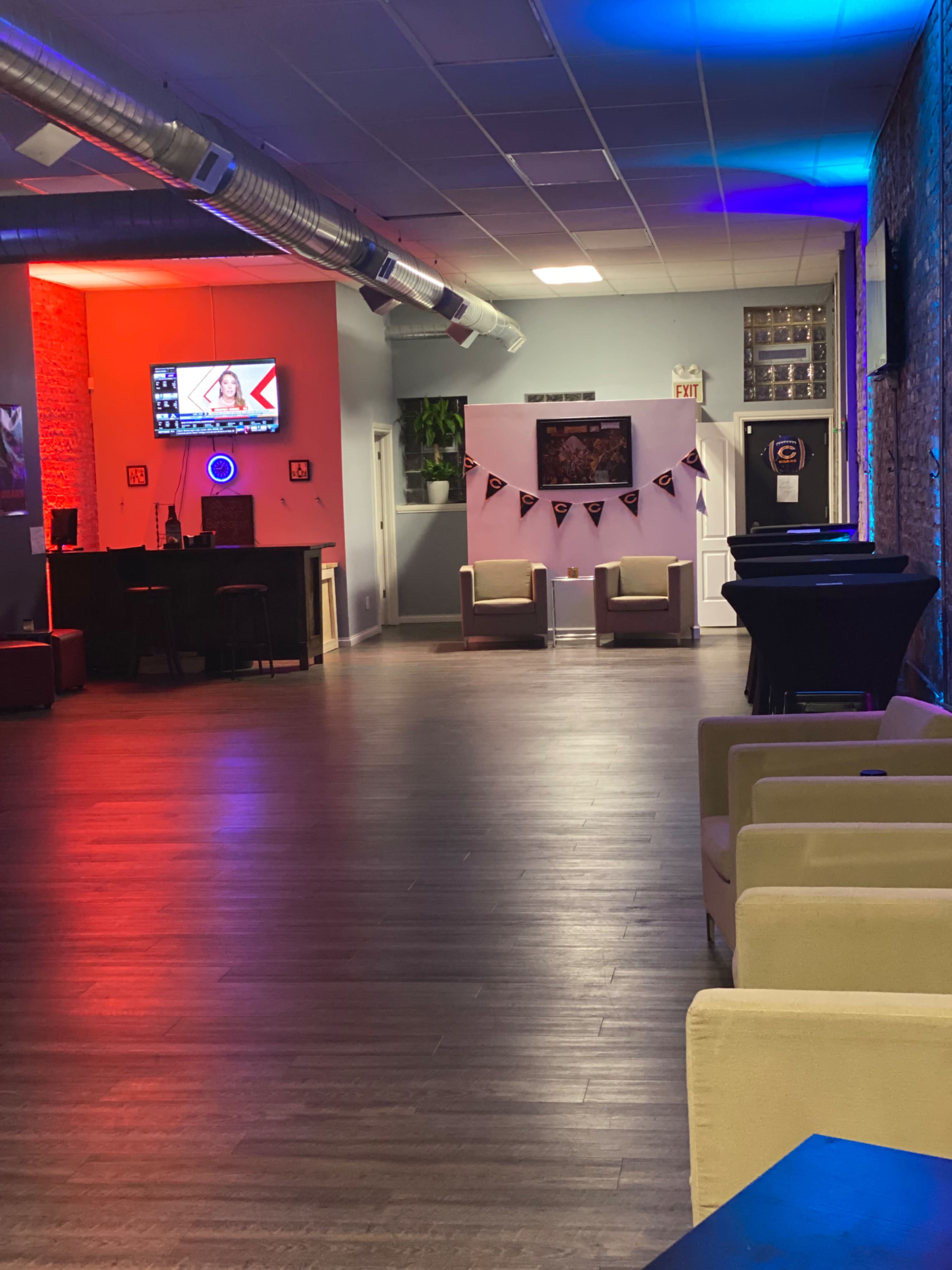 The image shows an interior space of a lounge or bar, featuring empty seating areas and a television displaying a program along one wall, with colorful lighting and decorations.