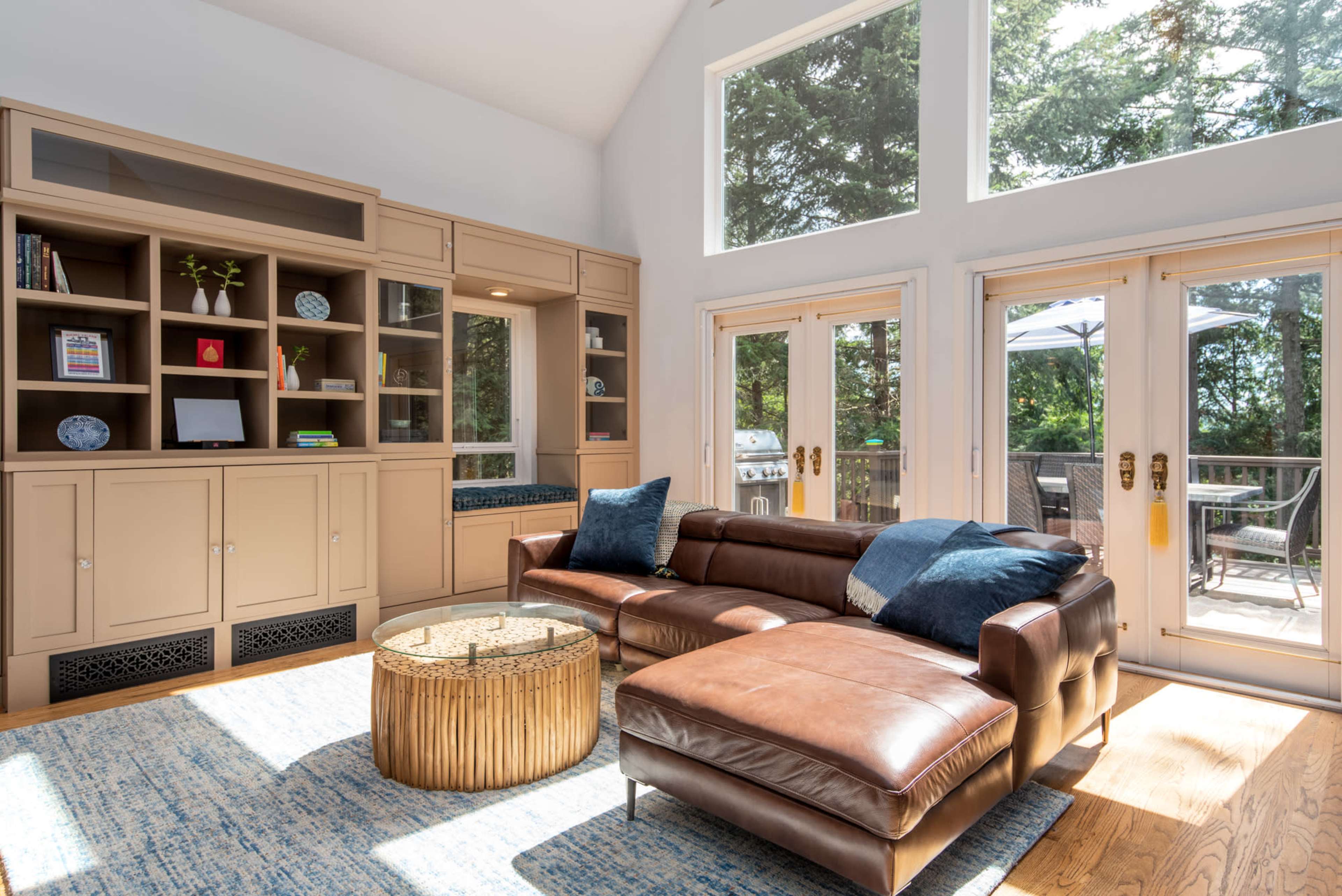 A sunlit living room features a brown leather sectional sofa, a circular gold coffee table, built-in cabinetry, and large windows overlooking a deck.