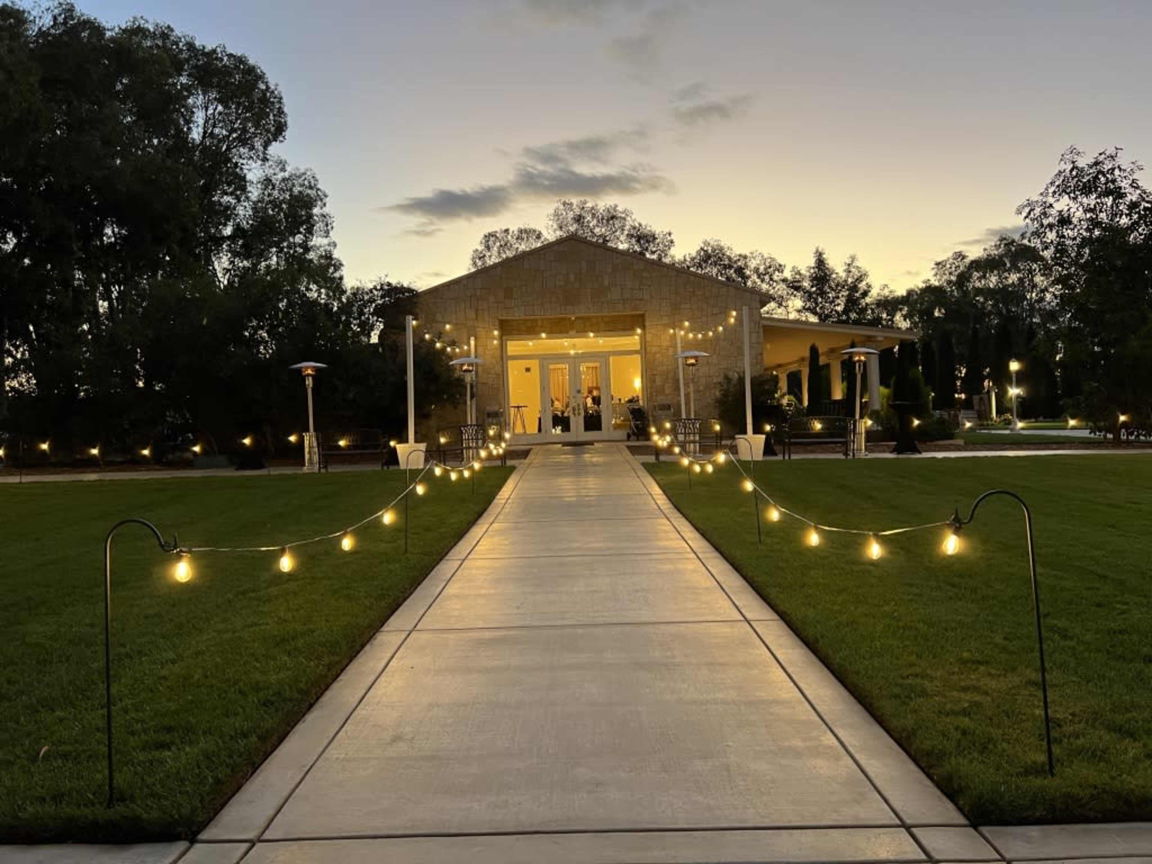A path illuminated by string lights leading to a well-lit building surrounded by greenery at dusk.