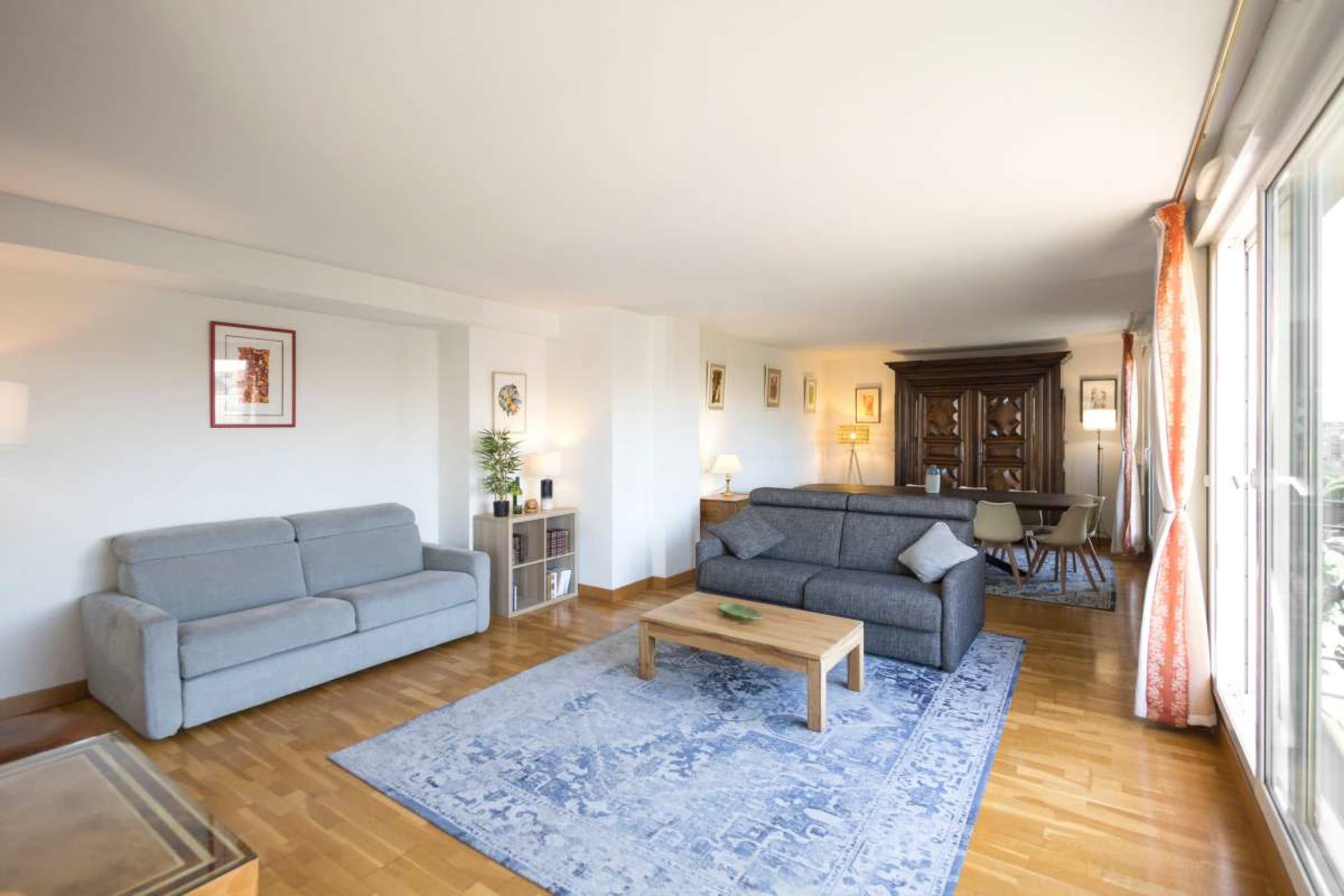 The image shows a bright living room with two gray sofas, a wooden coffee table on a blue area rug, and a dining area visible in the background.