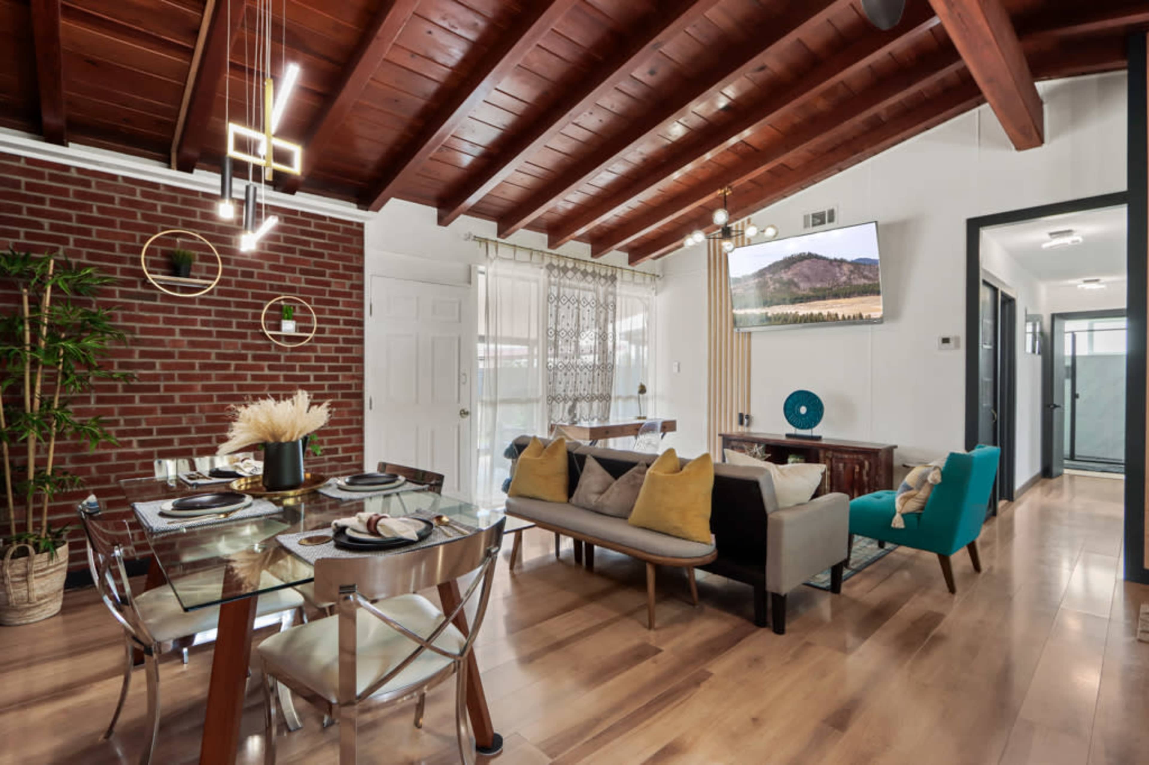 A modern dining area with a glass table, upholstered chairs, exposed wooden beams, and a brick accent wall.