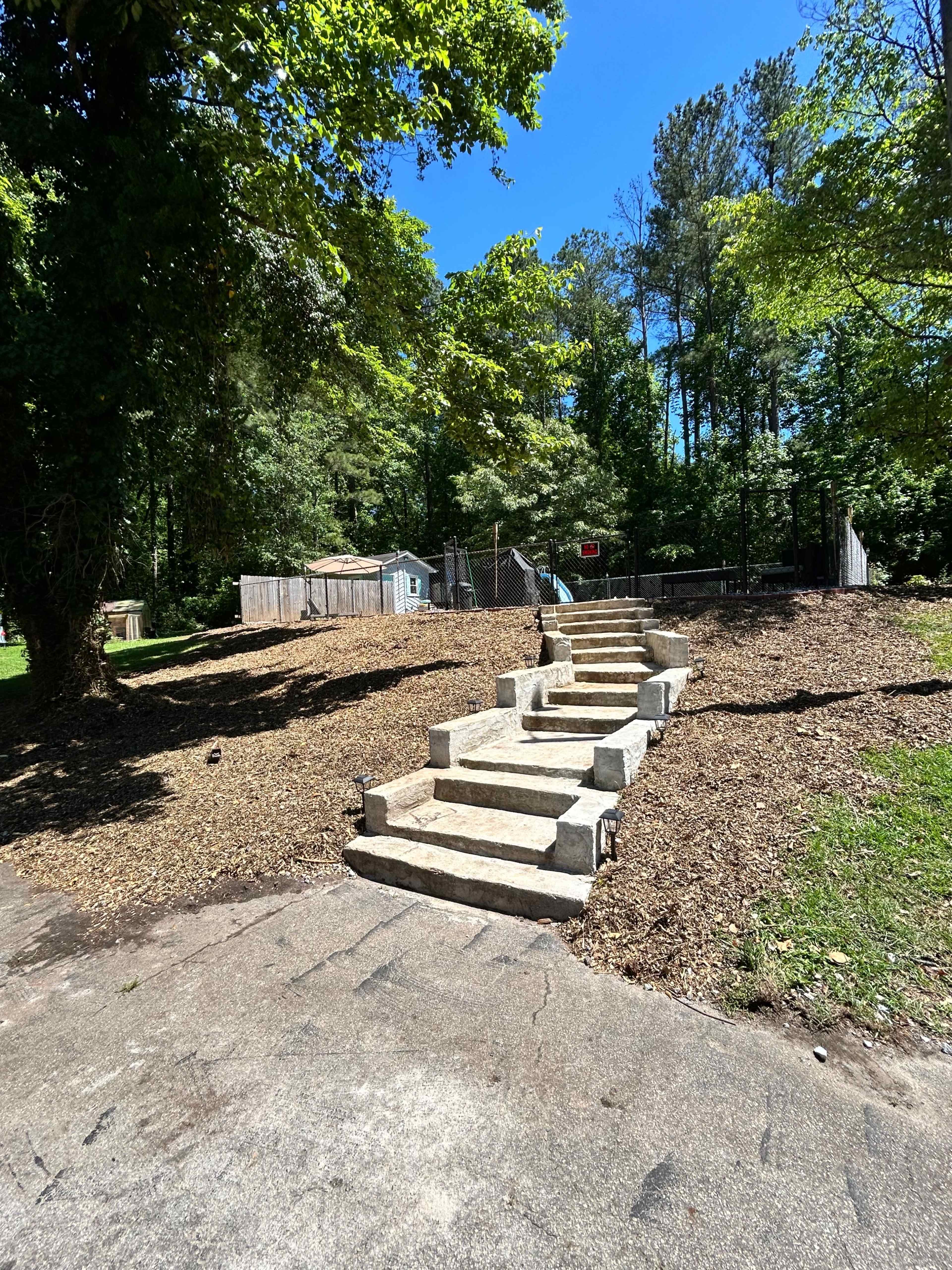 A set of stone steps leads up a grassy path toward a wooded area with some structures in the background.