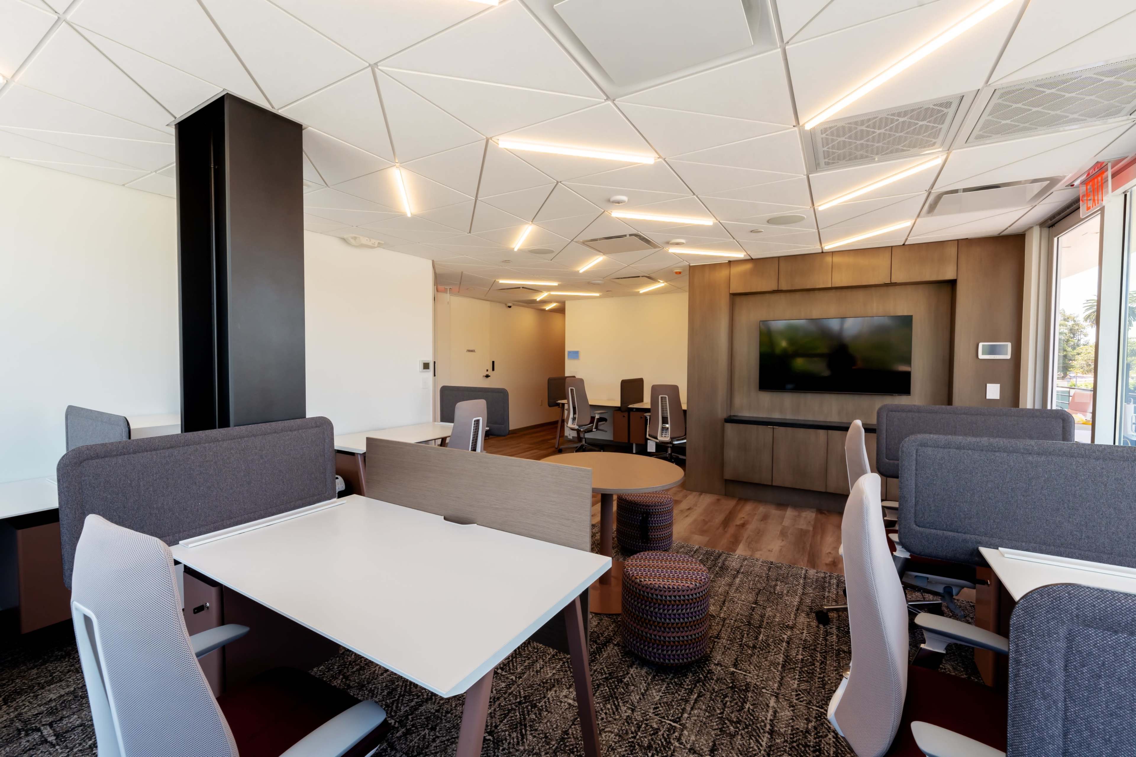A modern office space with individual workstations, lounge seating, and a wall-mounted television.
