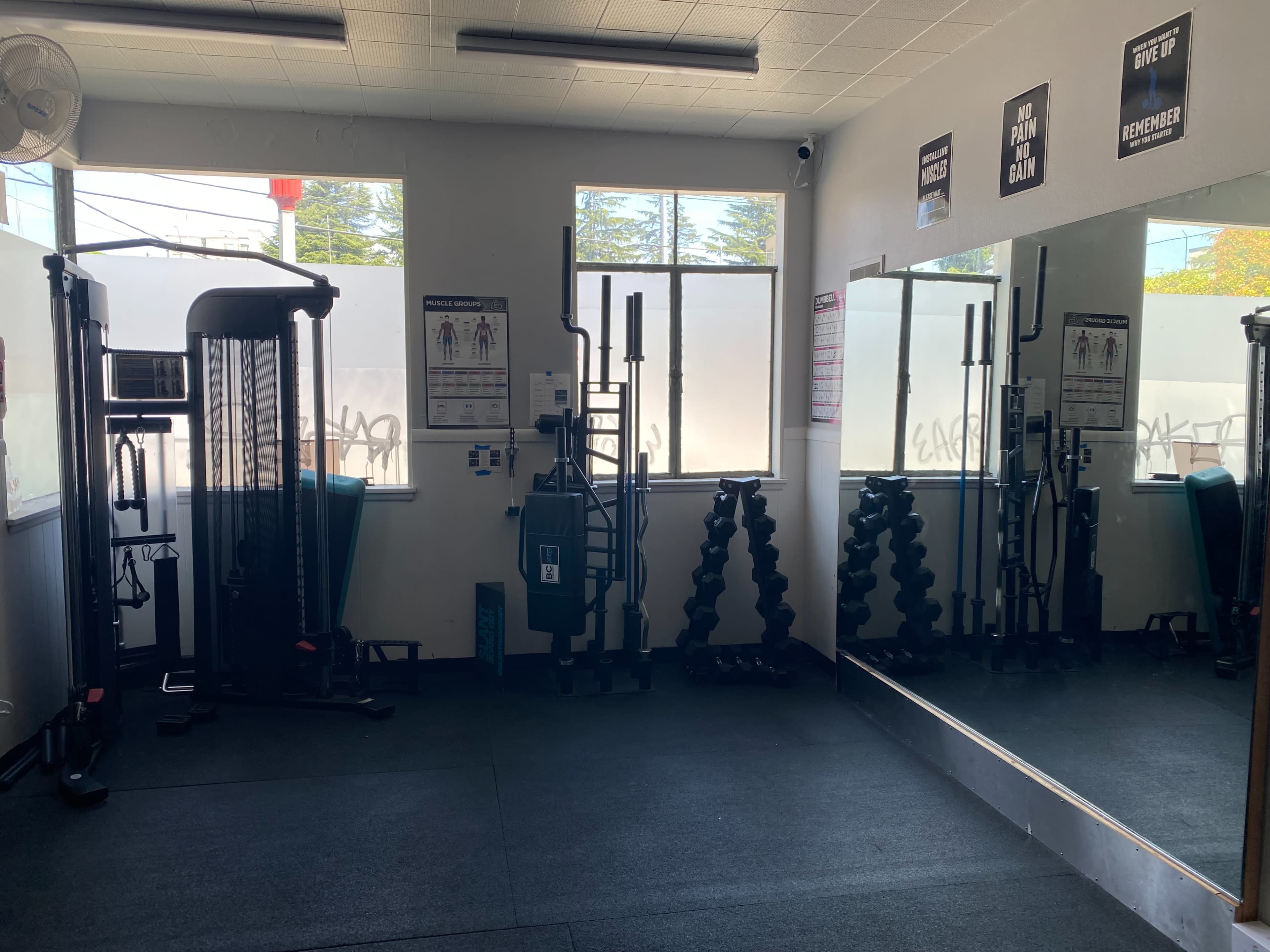 A gym interior with exercise equipment, including a cable machine, dumbbells, and mirrors on the walls.