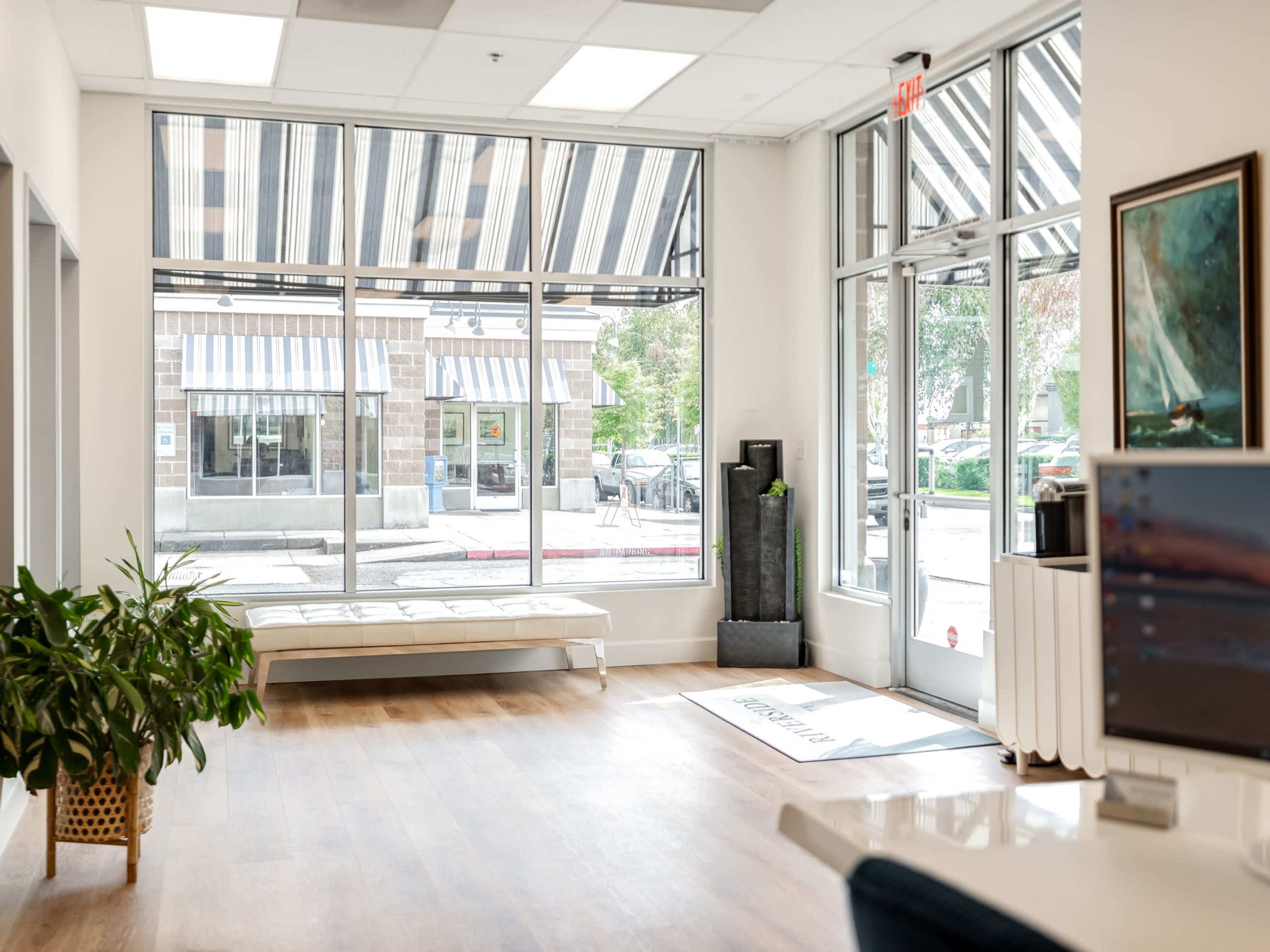 The image depicts a bright, modern interior with large windows, a bench along one wall, and a potted plant near the entrance.