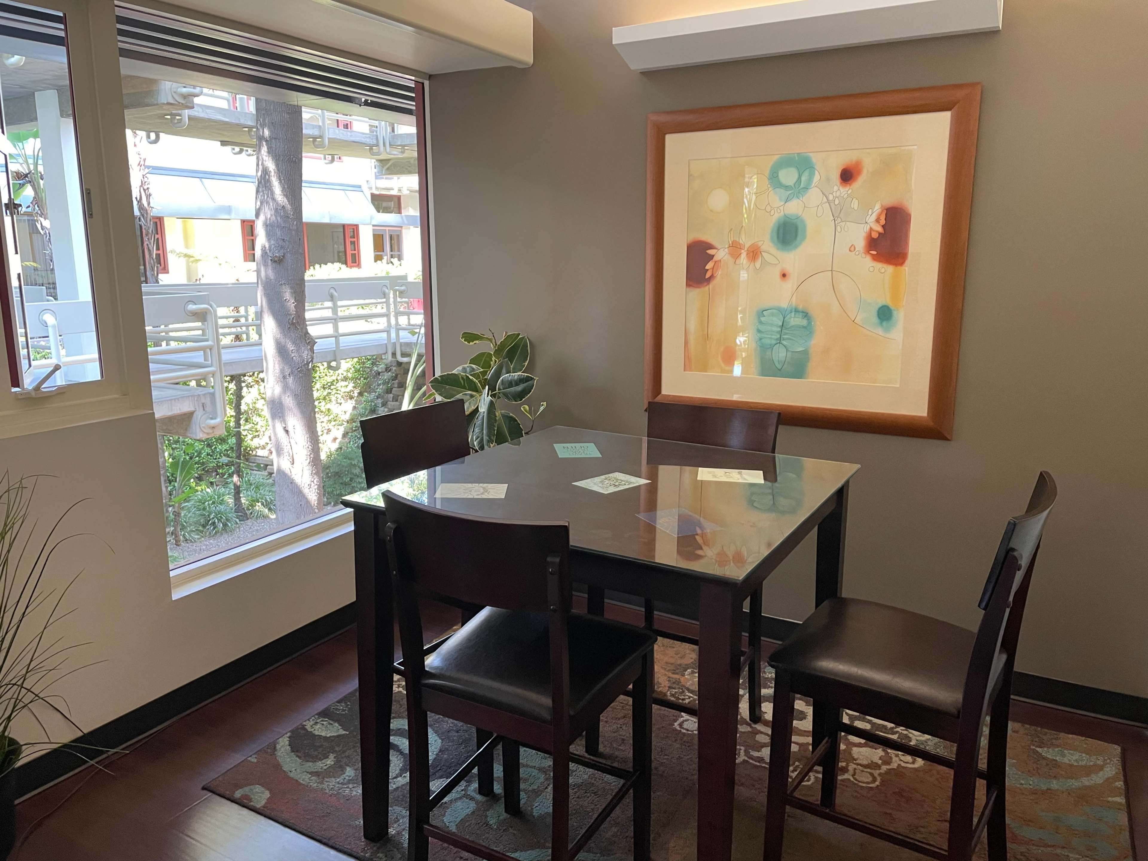 A dining area features a glass table surrounded by four chairs, with a framed artwork on the wall and a view of greenery outside through a large window.