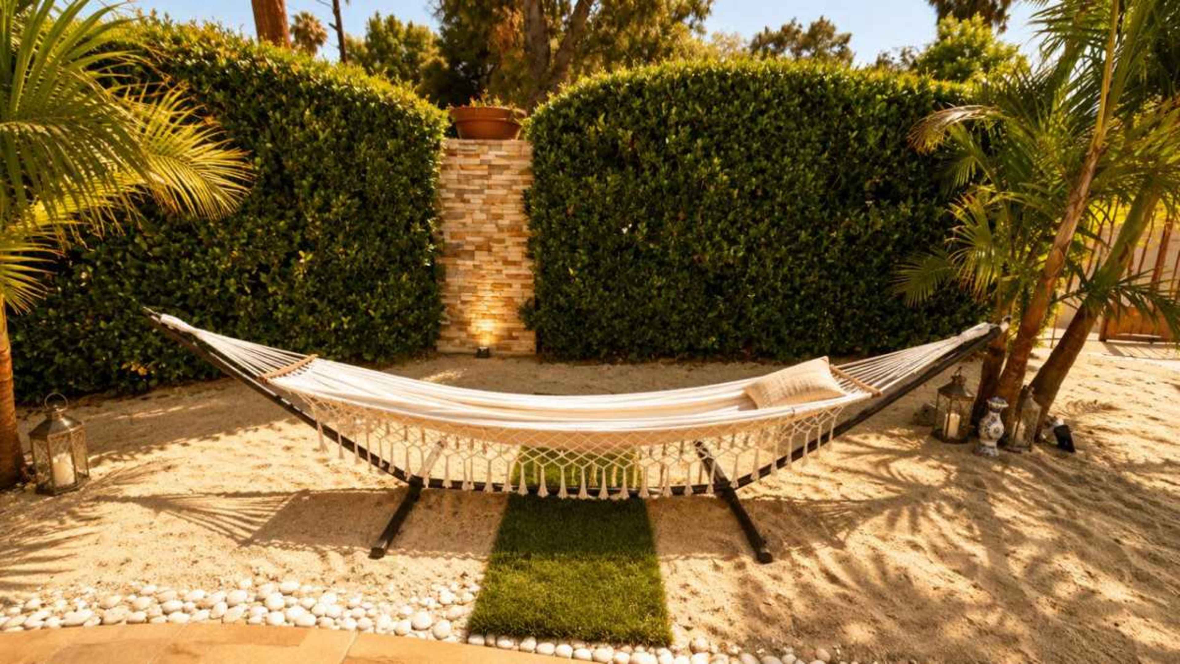A hammock is suspended between two palm trees in a sandy outdoor space surrounded by tall green hedges.