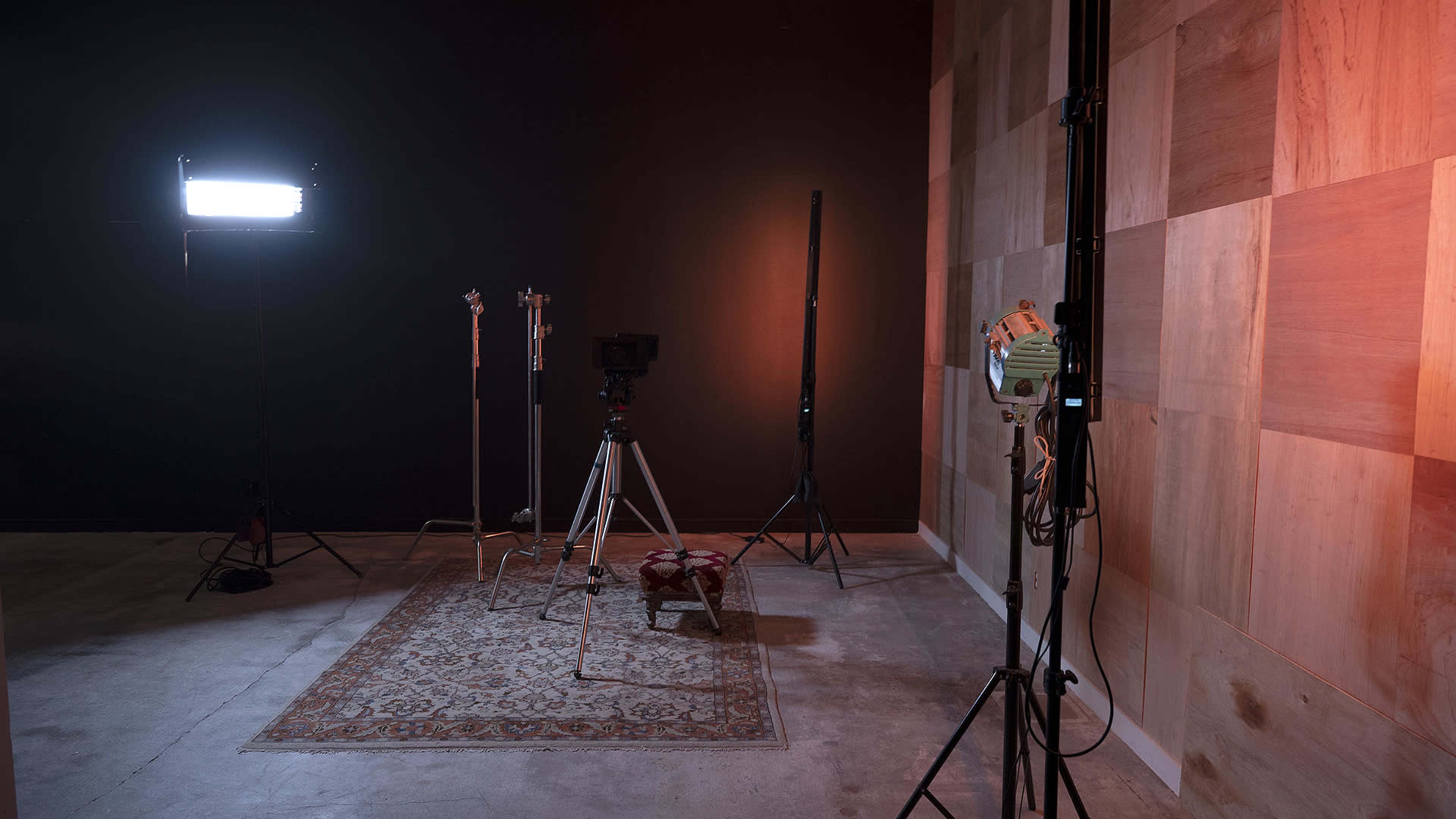 An empty studio space with lighting equipment, a camera on a tripod, and a patterned rug on the floor against a wooden-paneled wall.
