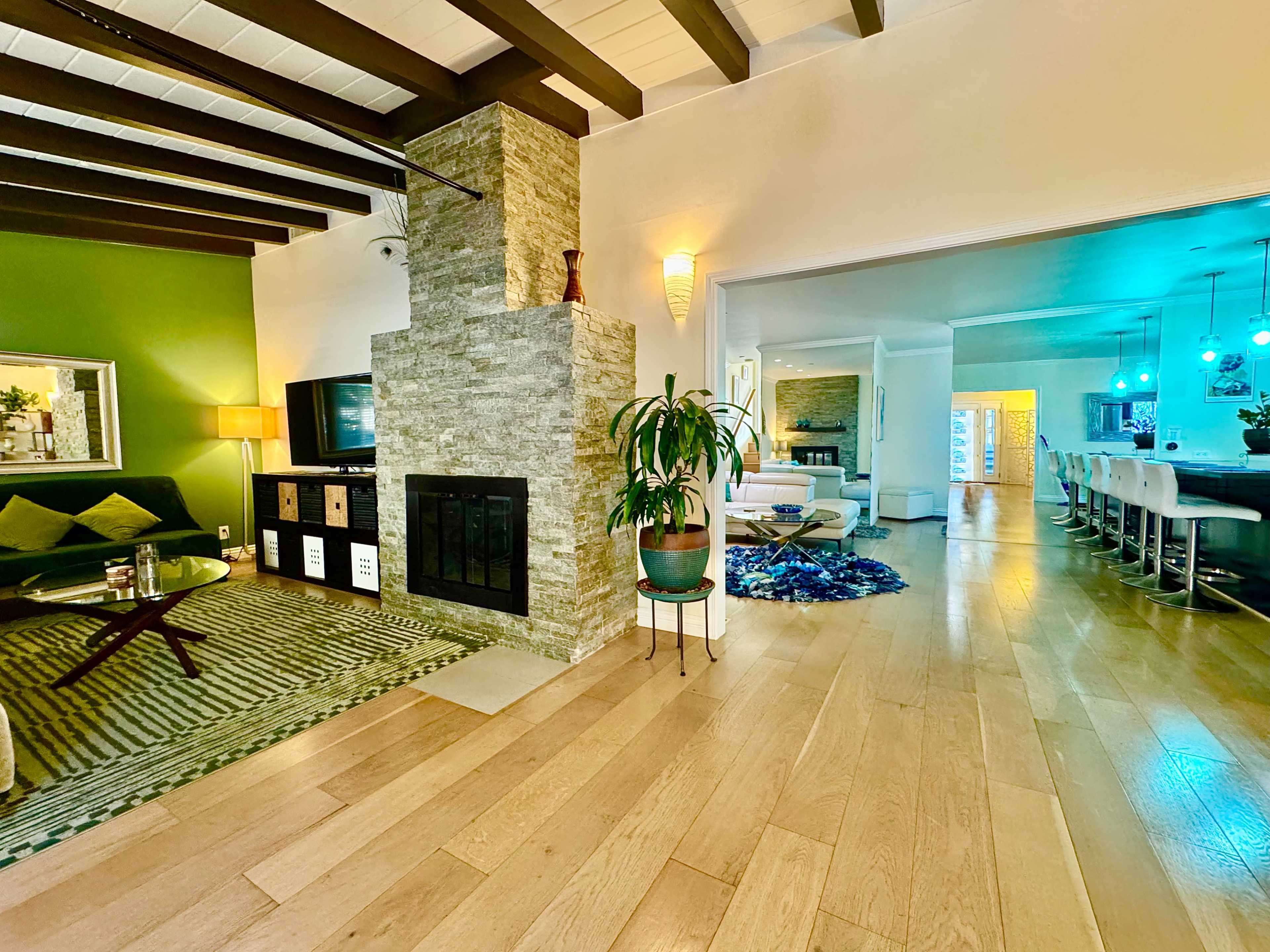 The image shows a spacious living area with a stone fireplace, green accent wall, and an open layout leading to a dining area with bar stools.