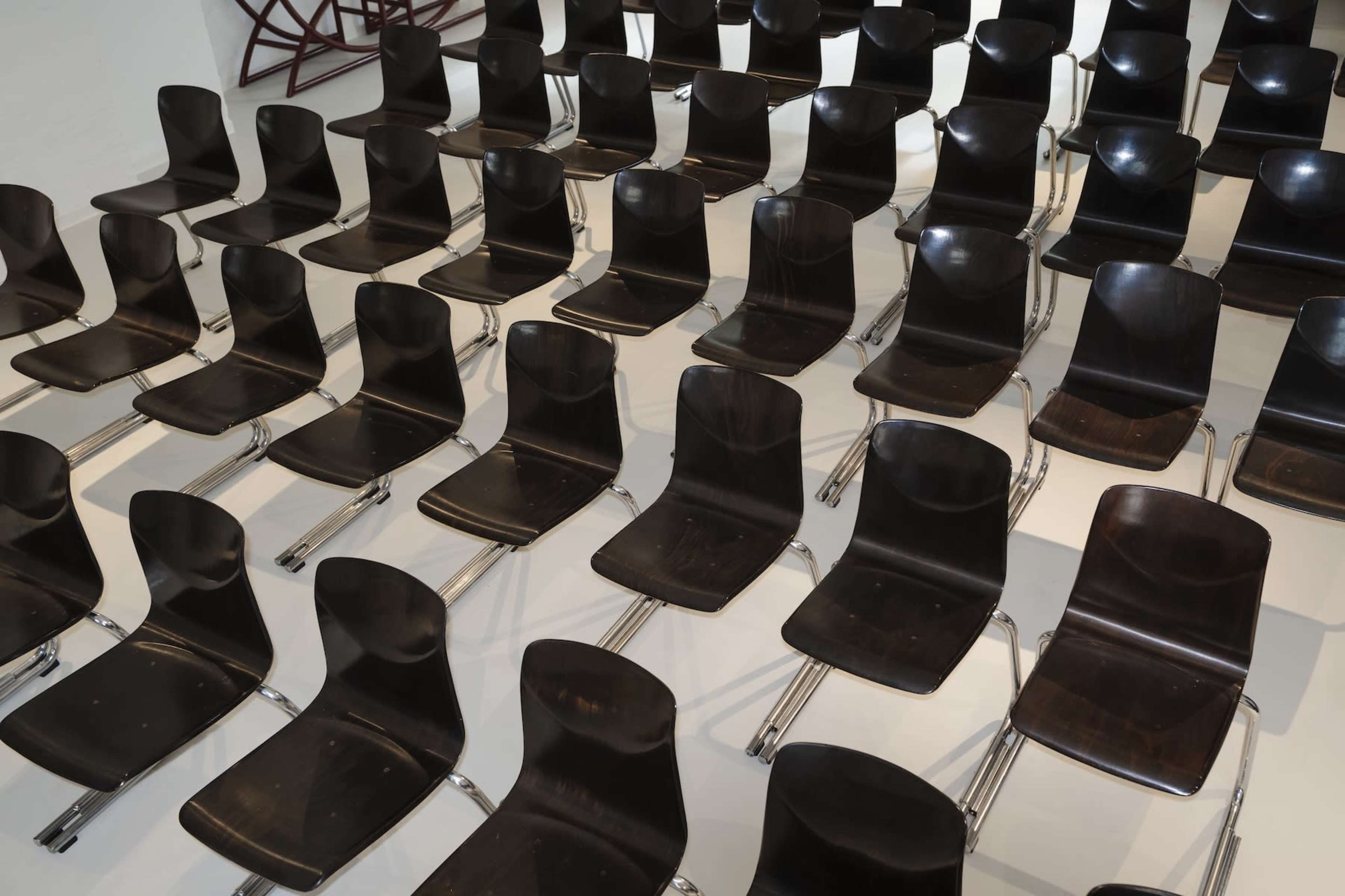 The image shows a high-angle view of numerous black wooden chairs arranged neatly in rows on a white floor.