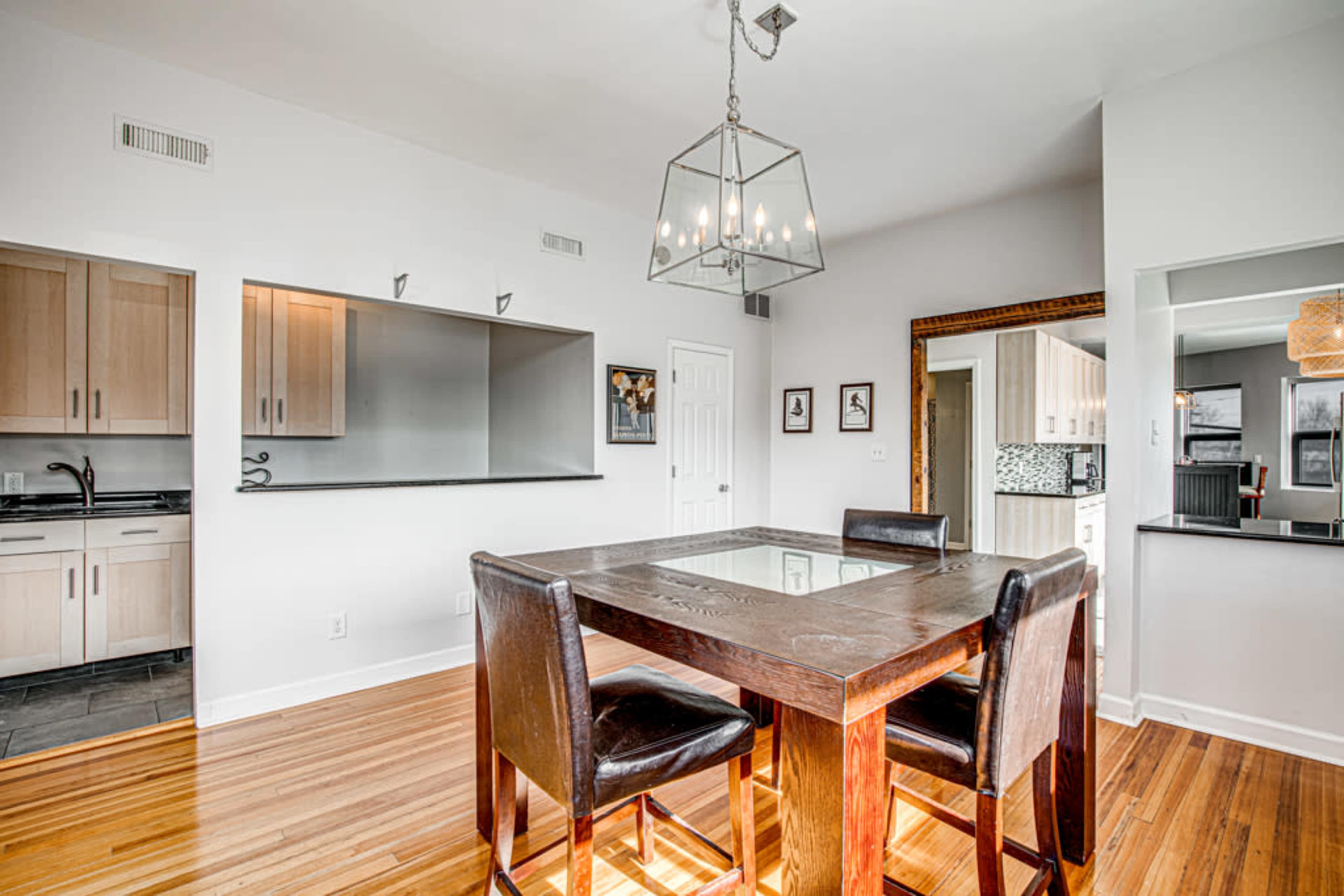 A modern kitchen and dining area features a wooden table with dark chairs, illuminated by a geometric ceiling light, with minimalist cabinetry and a large opening to adjacent rooms.