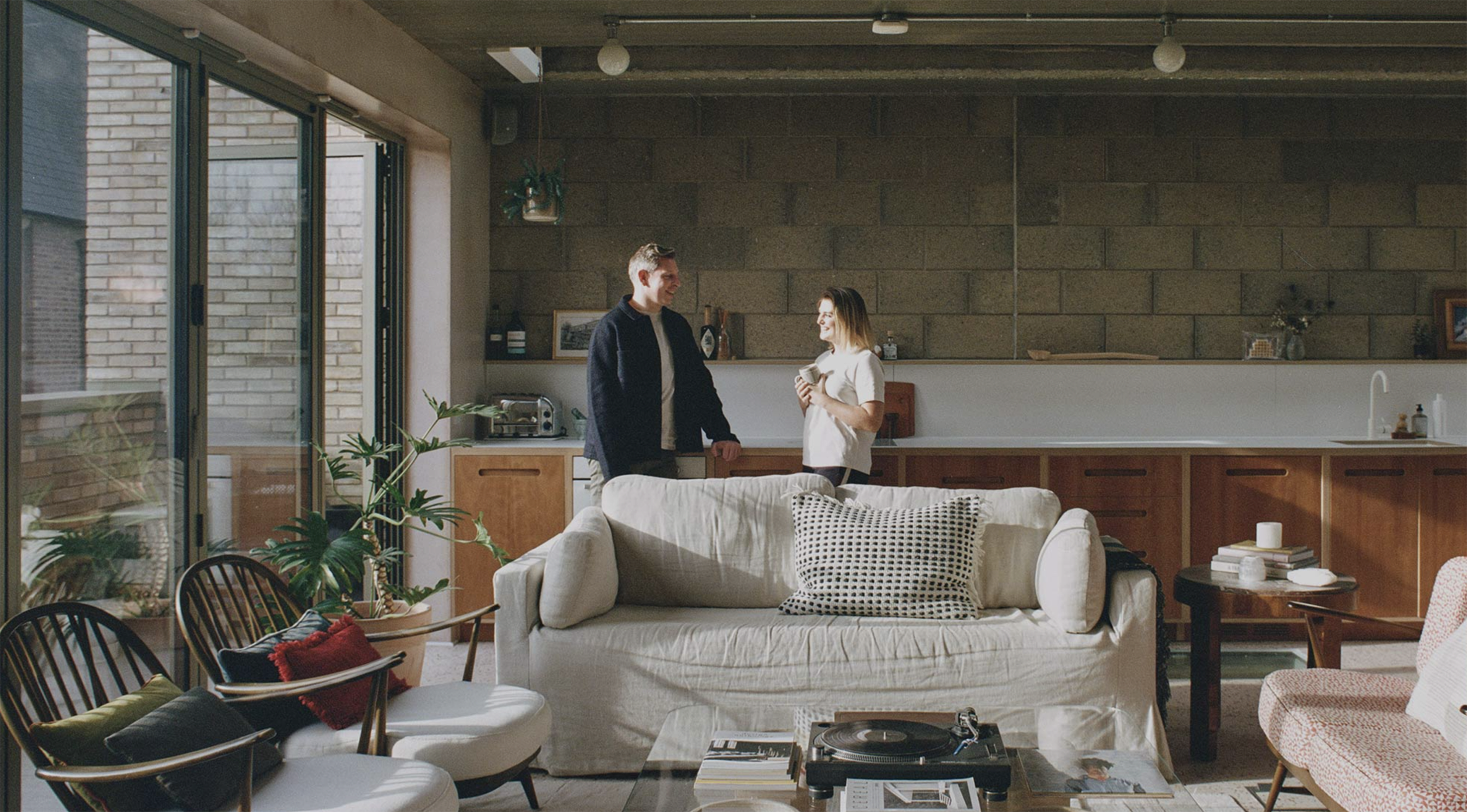 A man and a woman stand in a modern kitchen area, engaged in conversation, with a spacious living room furnished with a sofa, seating, and houseplants visible in the background.