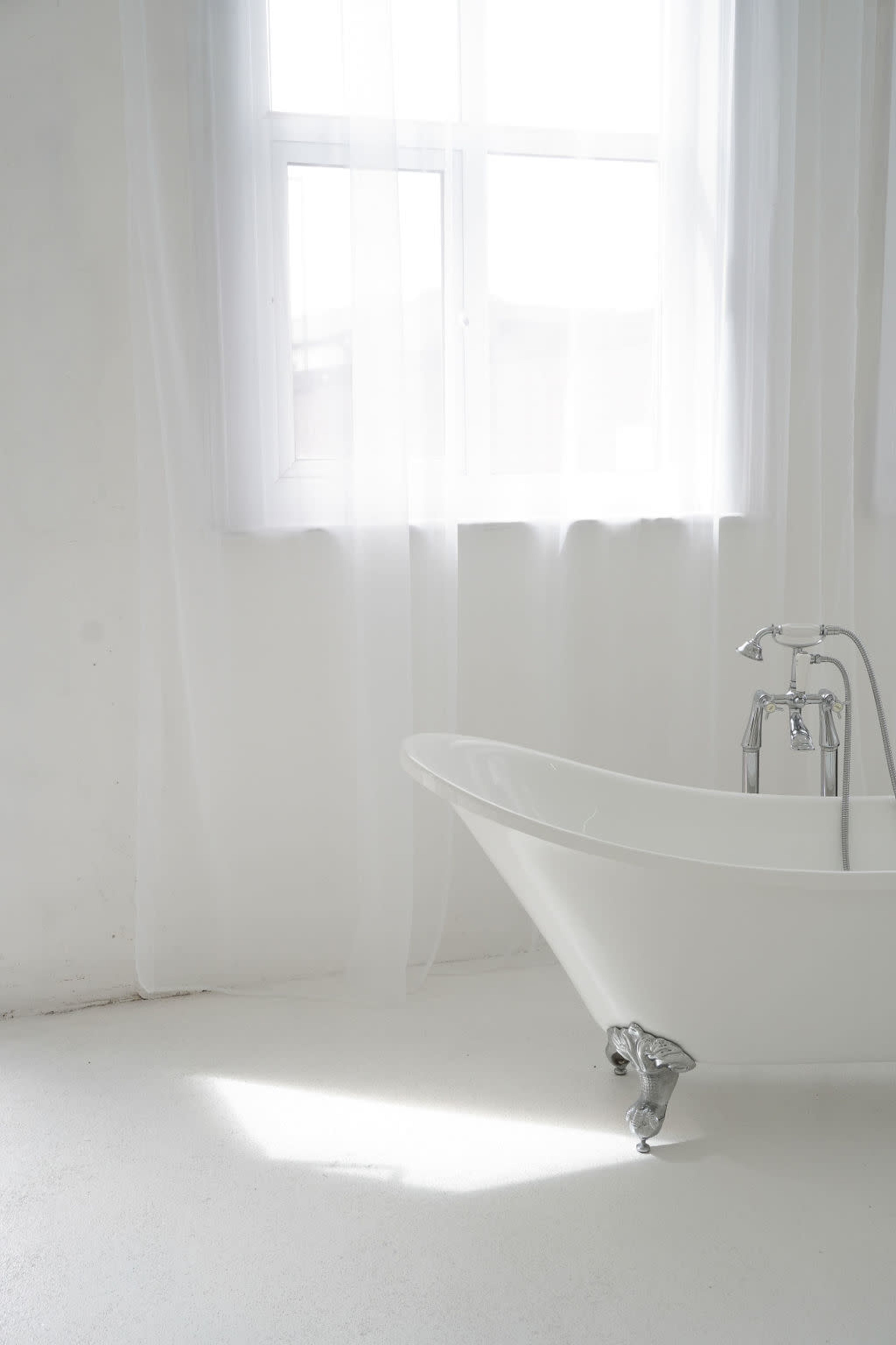 A freestanding white bathtub is positioned near a window with sheer white curtains in a bright, minimalistic room.
