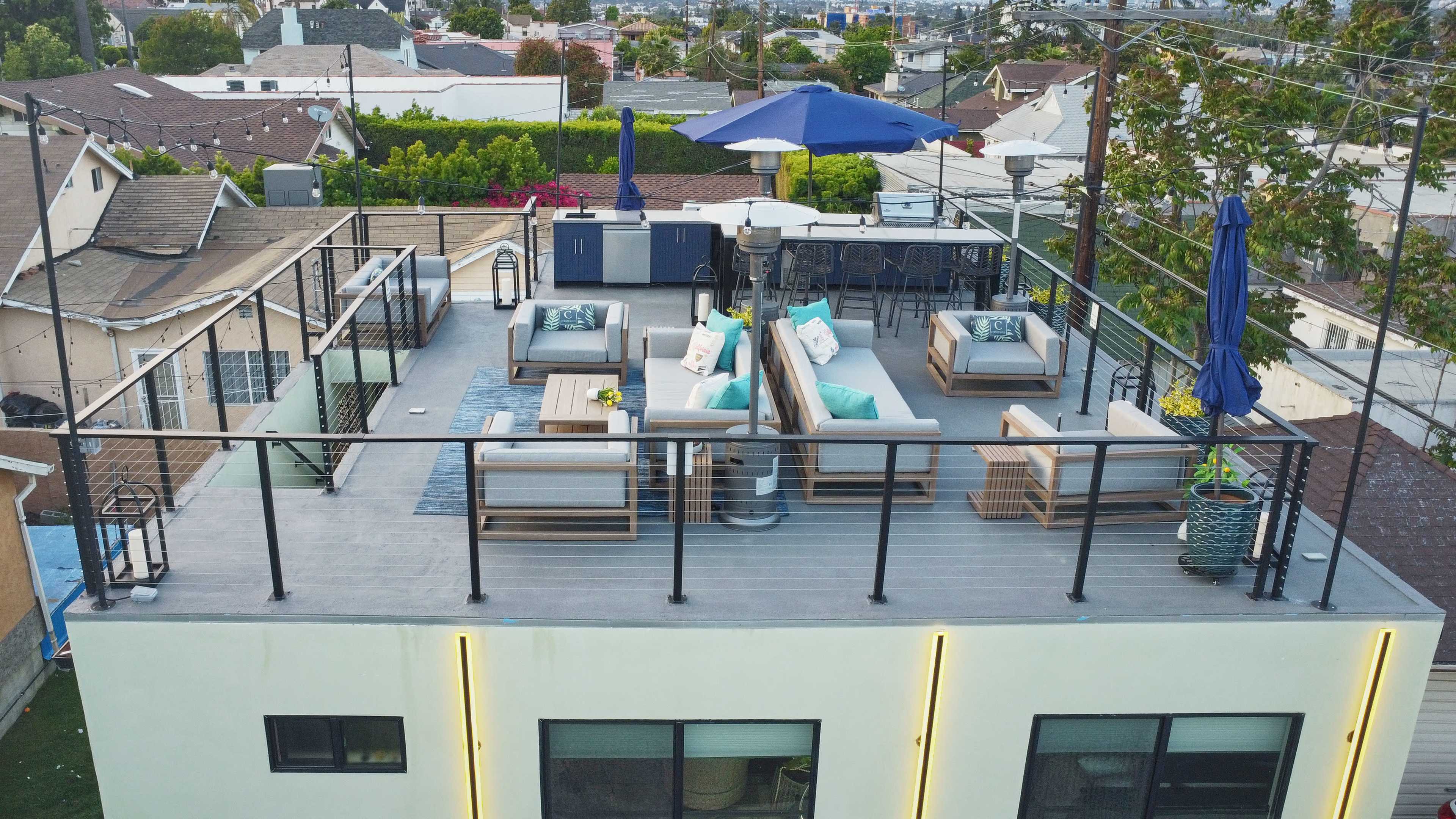 The image shows a rooftop terrace furnished with seating areas, umbrellas, and a dining setup, surrounded by residential rooftops.
