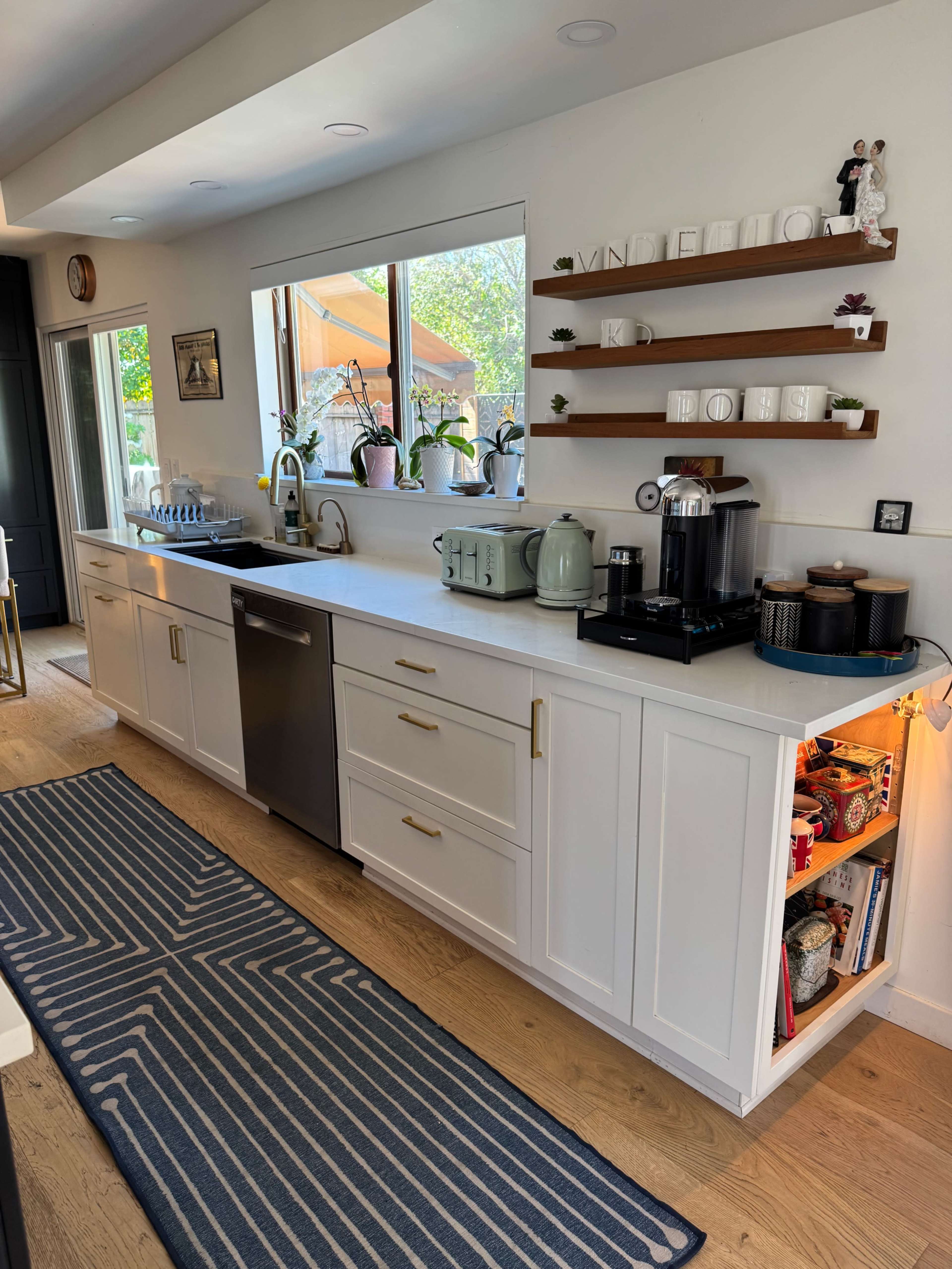 The image shows a modern kitchen with white cabinetry, a stainless steel dishwasher, and a countertop coffee station, featuring a mix of utensils and decorative items on shelves.