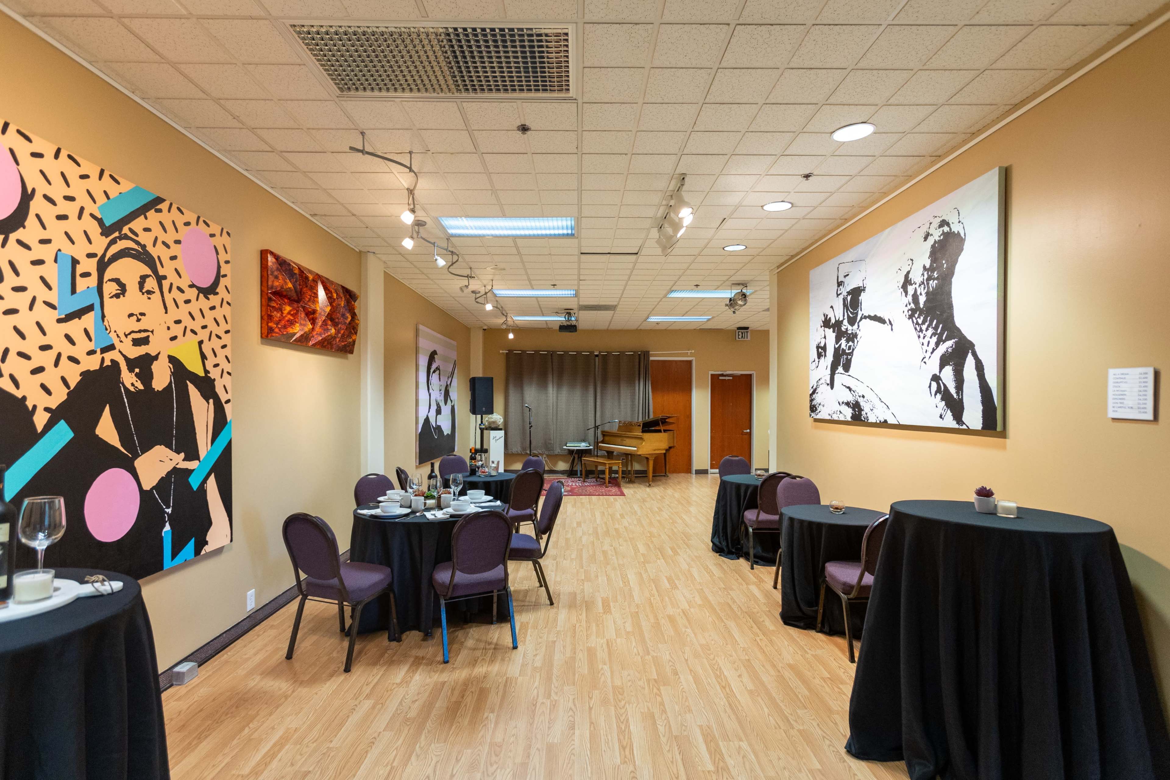 A well-lit event space with tables set for dining, featuring artwork on the walls and a stage area with a piano at the far end.