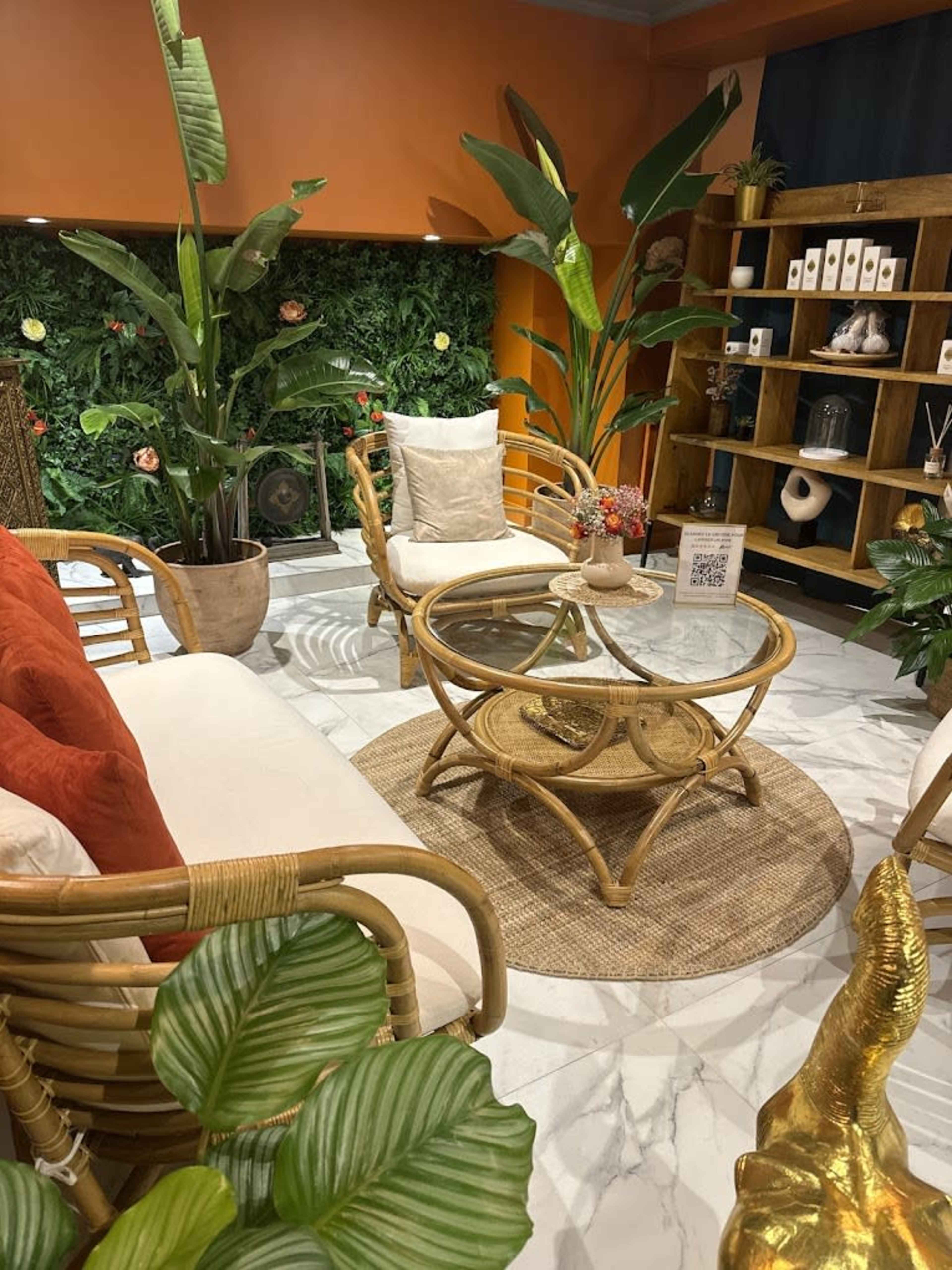 The image shows a cozy seating area with rattan furniture, a round glass coffee table, and large indoor plants, set against a green foliage backdrop and orange walls.