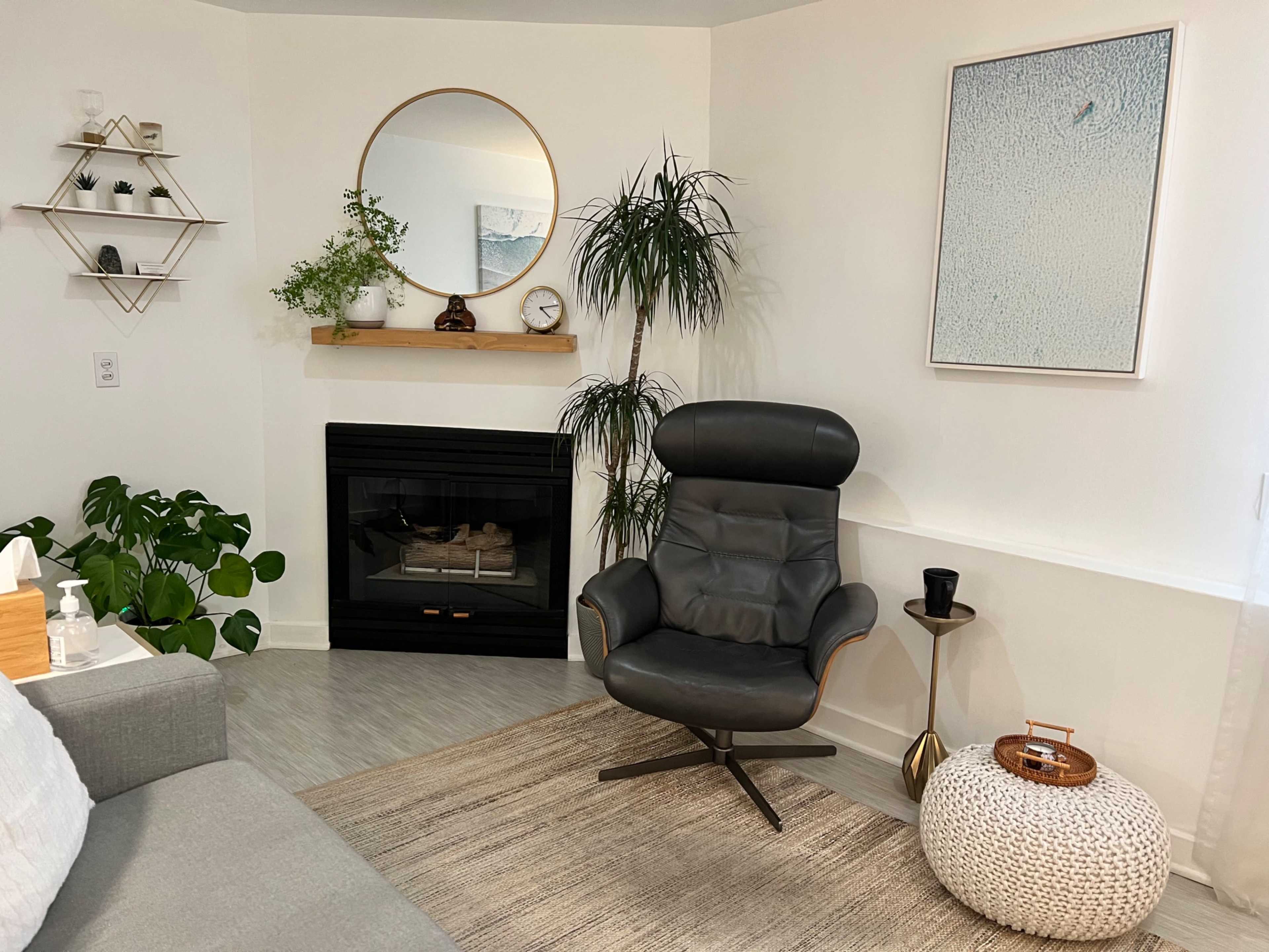 A modern living room features a cozy gray sofa, a black leather chair, a round mirror, and a minimalist decor with plants and a fireplace.