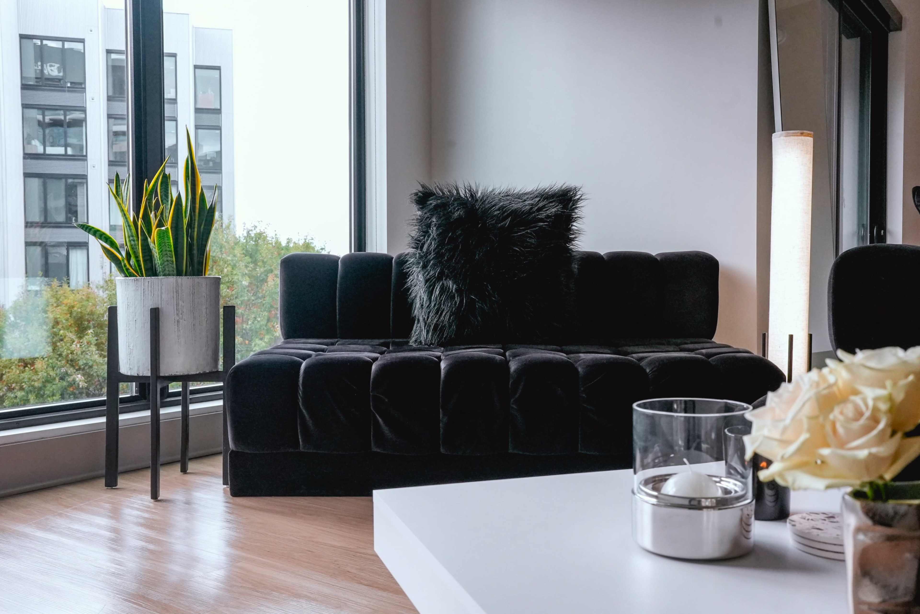A black velvet couch with a plush pillow sits beside a large window, overlooking greenery, with a potted plant and a glass candle holder on a white table.