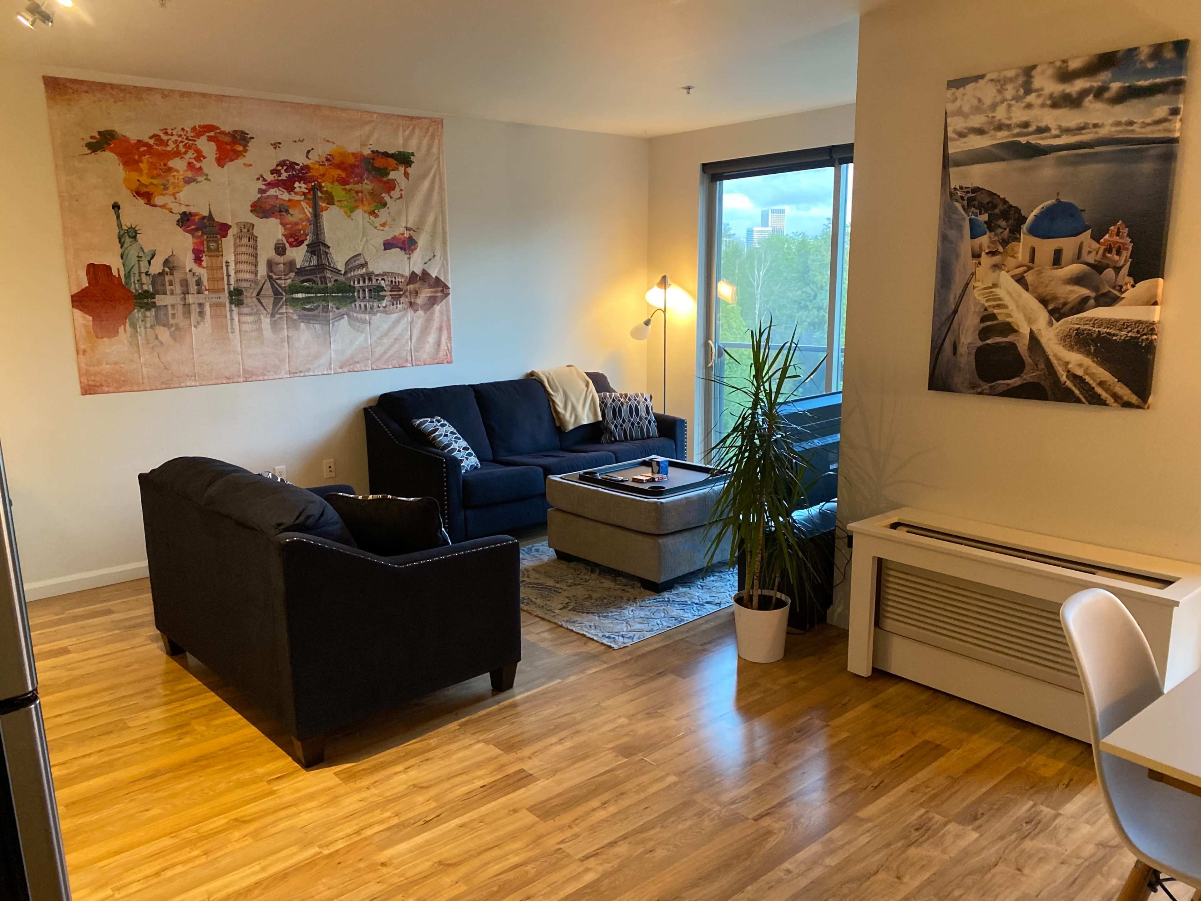 A modern living room featuring a map-themed tapestry on the wall, two black sofas, a coffee table, and large windows with natural light.
