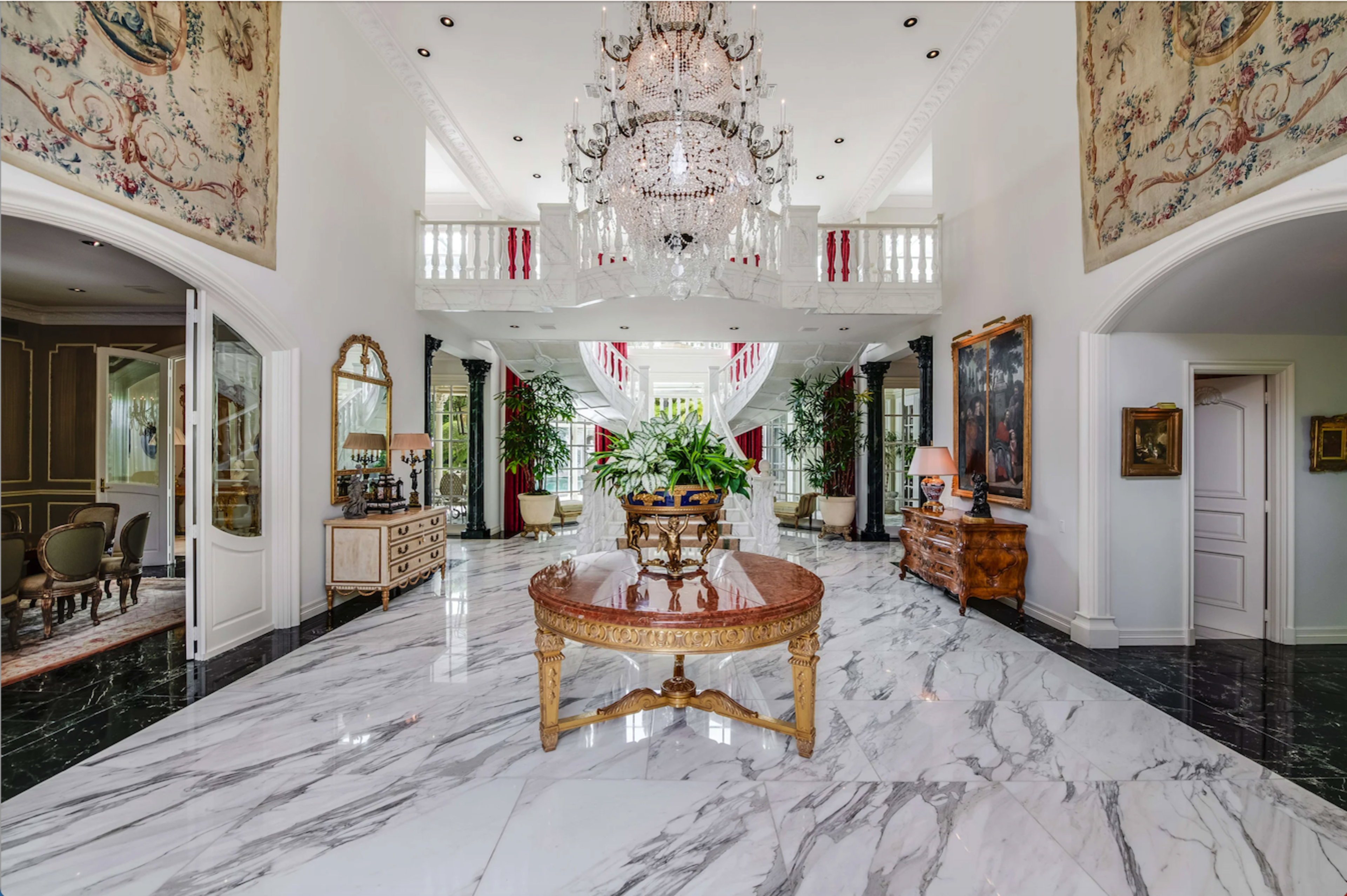 The image shows a grand foyer with a chandelier, marble flooring, and ornate decor, leading to a staircase and various rooms.