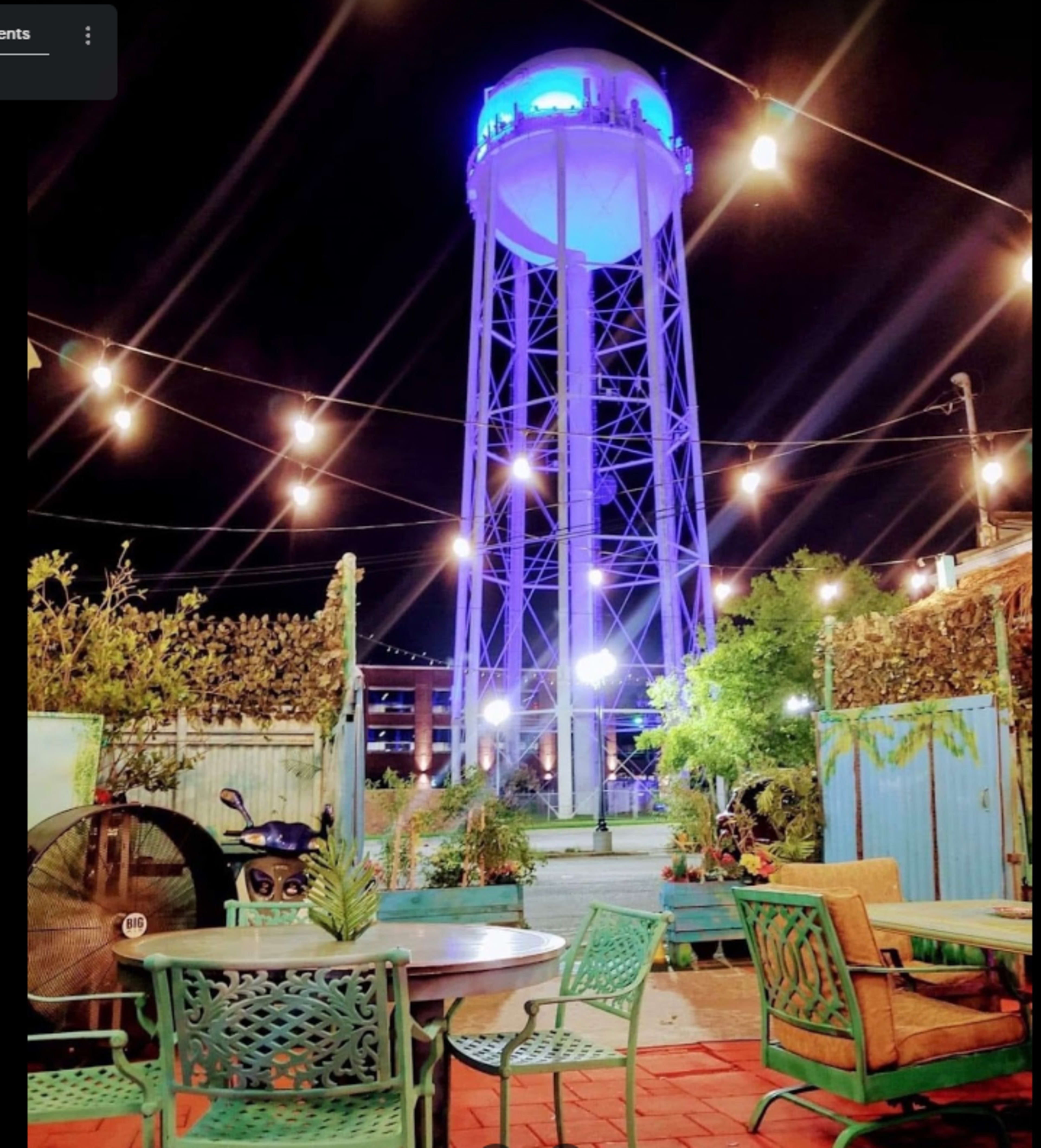 A brightly lit water tower stands illuminated in blue amidst outdoor seating and string lights in an urban courtyard at night.