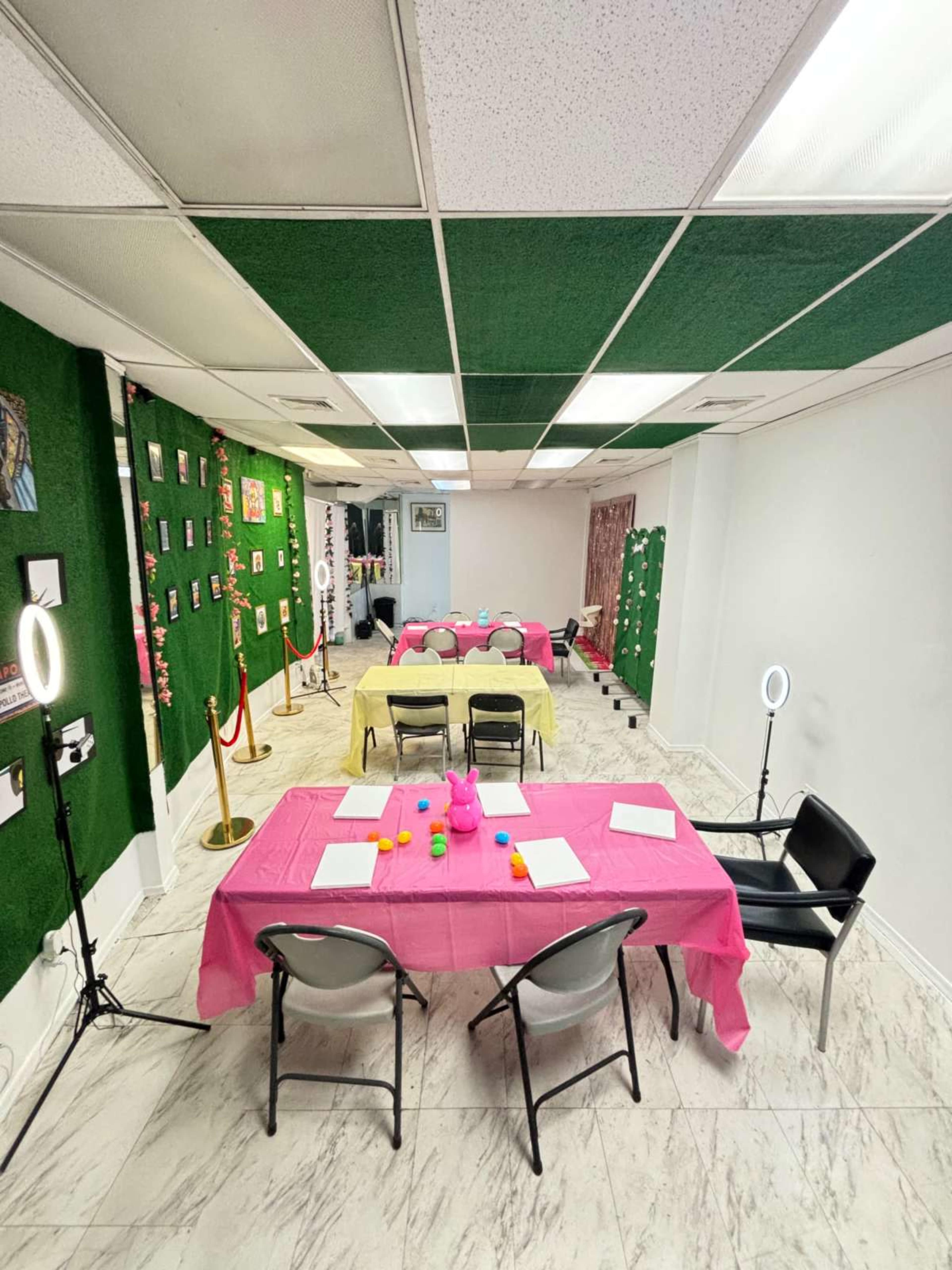 A large room set up with several tables covered in pink cloth, surrounded by chairs, decorated walls, and lighting equipment.
