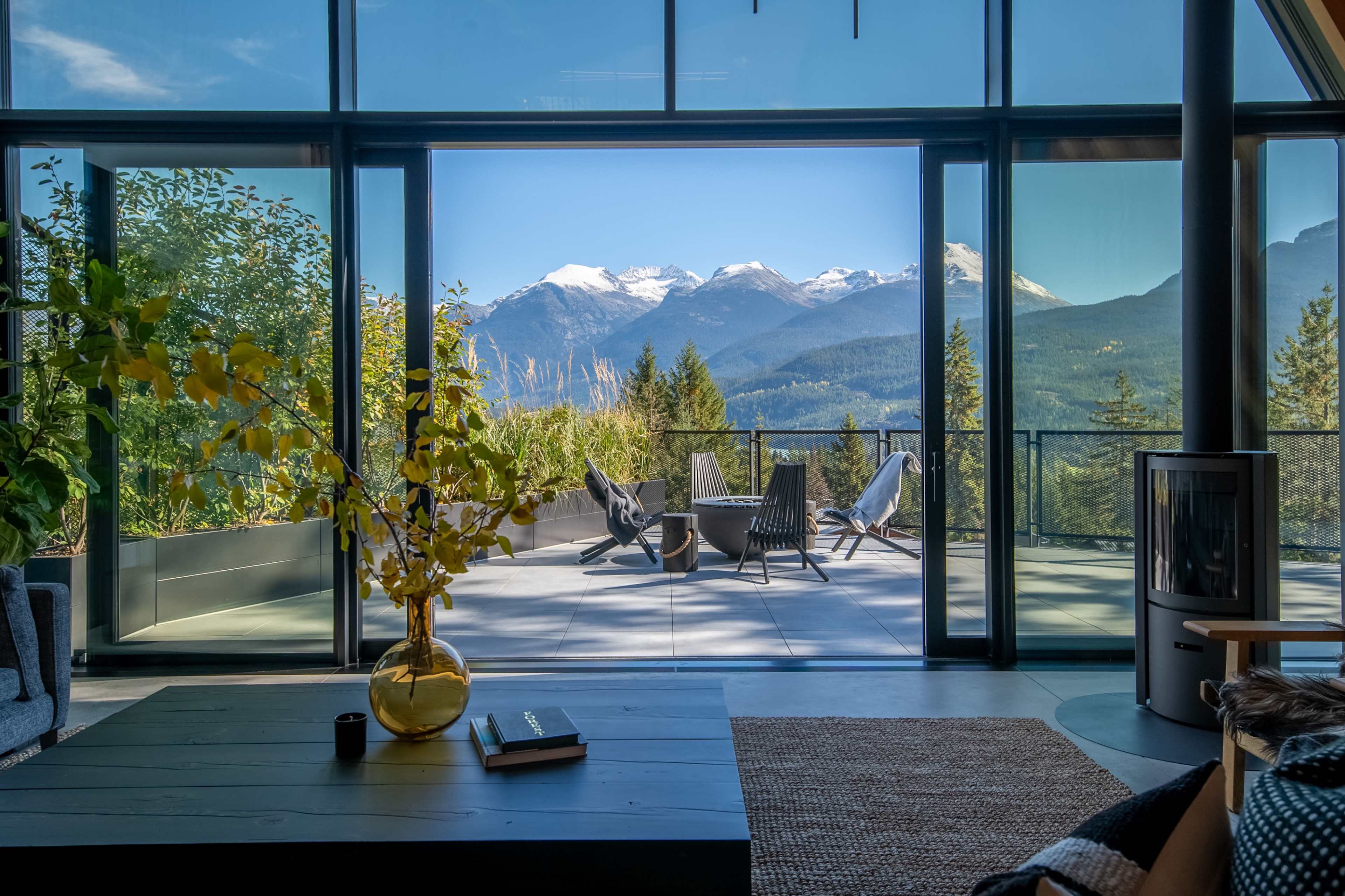 The image shows a modern living room with large glass doors leading to a terrace, offering a view of snow-capped mountains and a forested landscape.