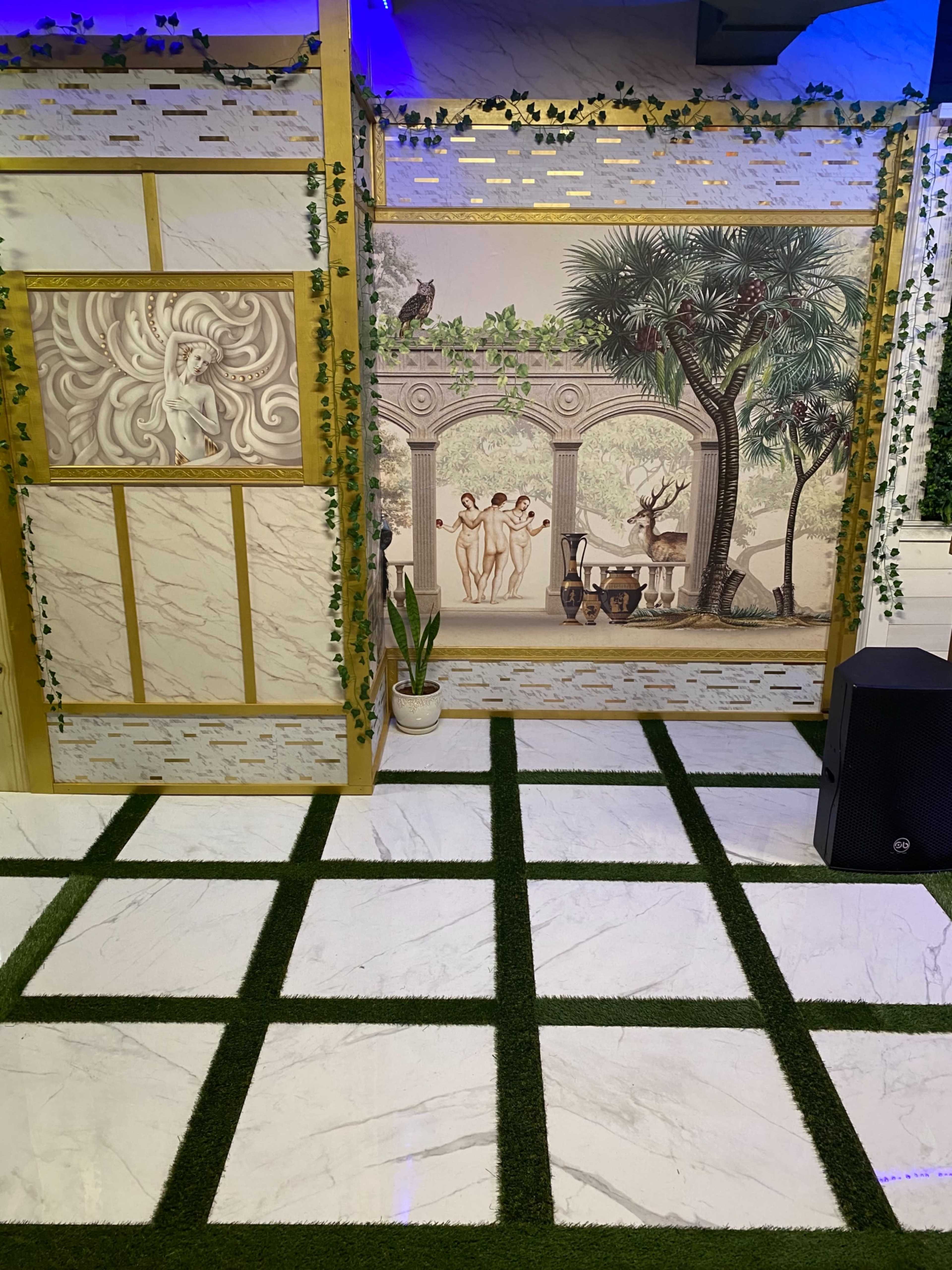 A decorated indoor space featuring a mural with people in a garden setting, surrounded by green artificial grass and a speaker.