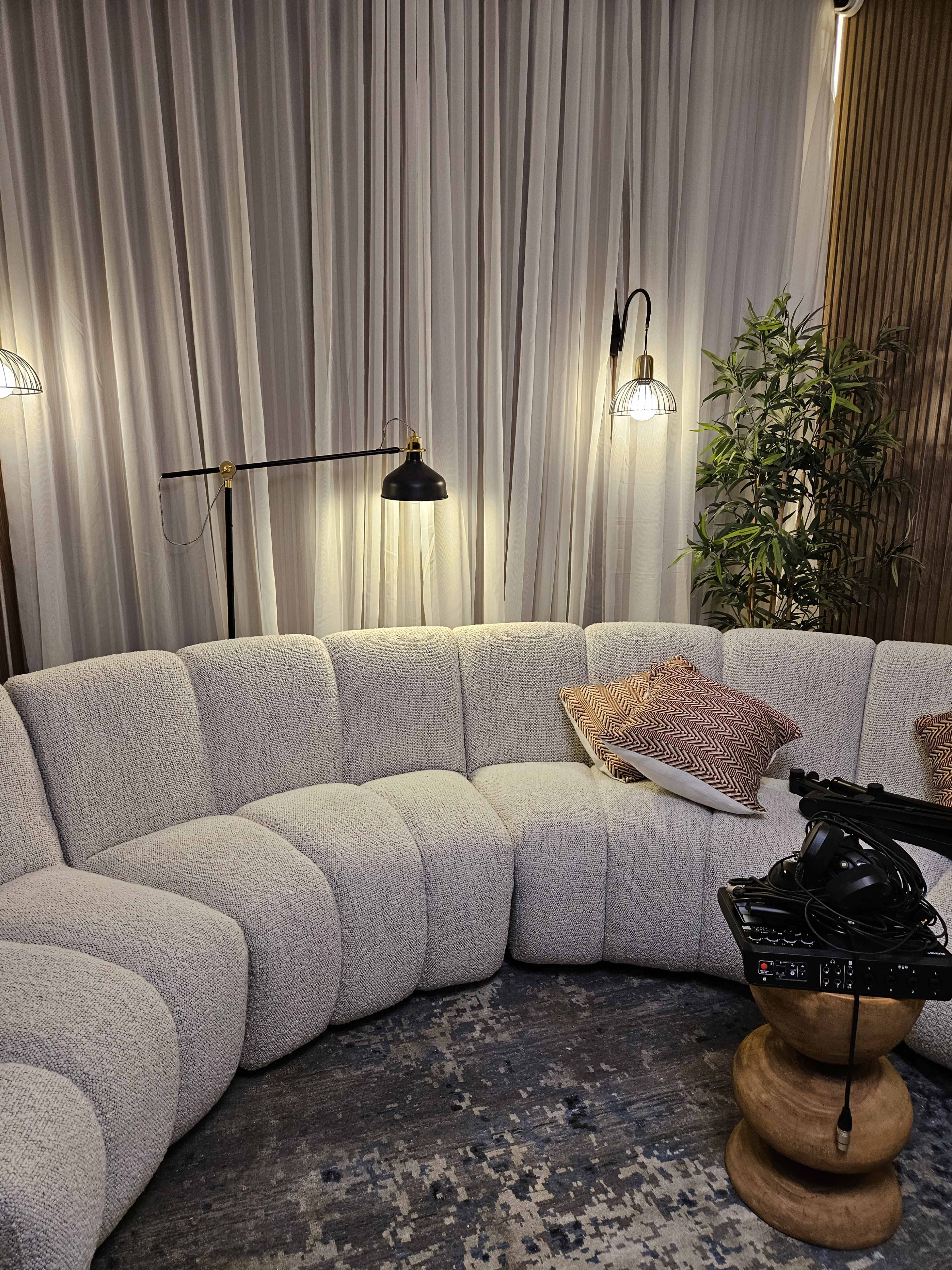 A curved, light-colored sofa is positioned in a cozy room illuminated by stylish lamps and accented with decorative pillows, alongside a small wooden table holding a record player and a plant.