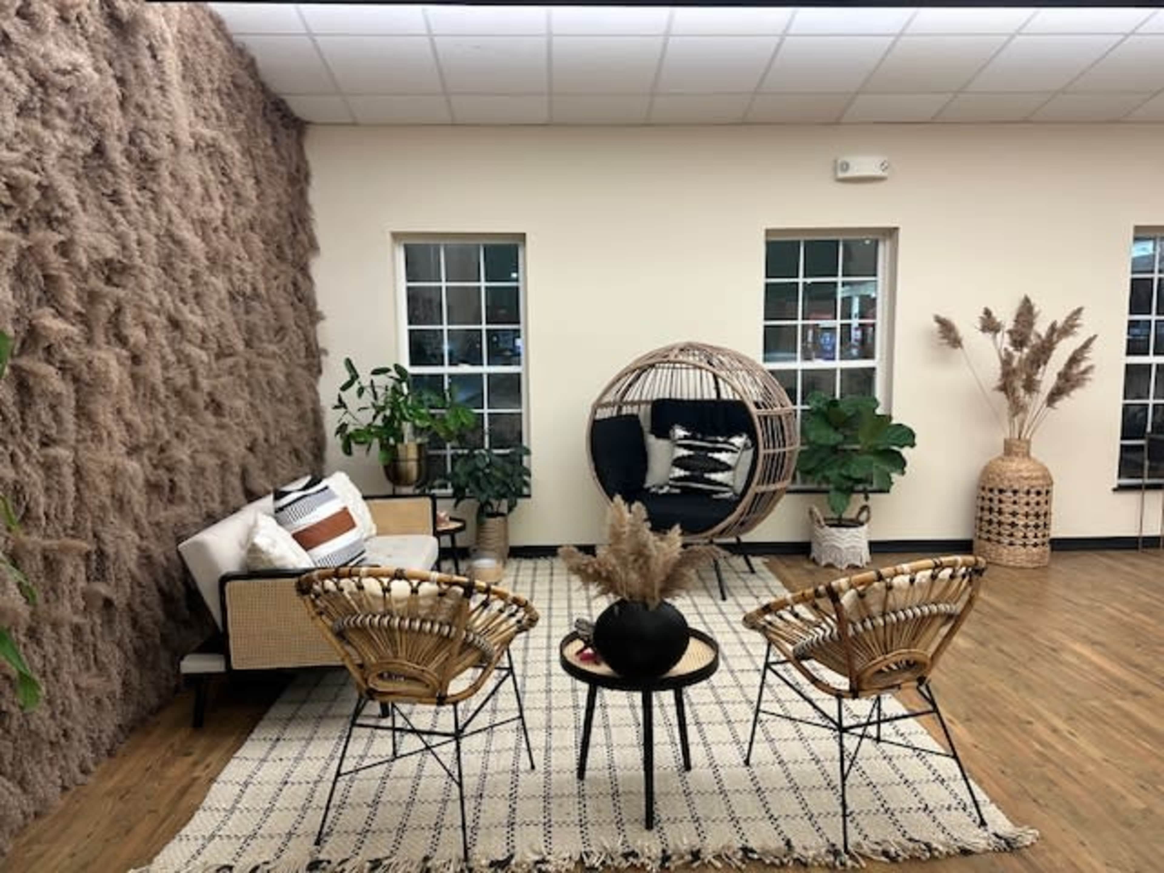 A cozy interior space featuring a hanging chair, a sofa, two woven chairs, and several plants arranged around a patterned rug on the wooden floor.