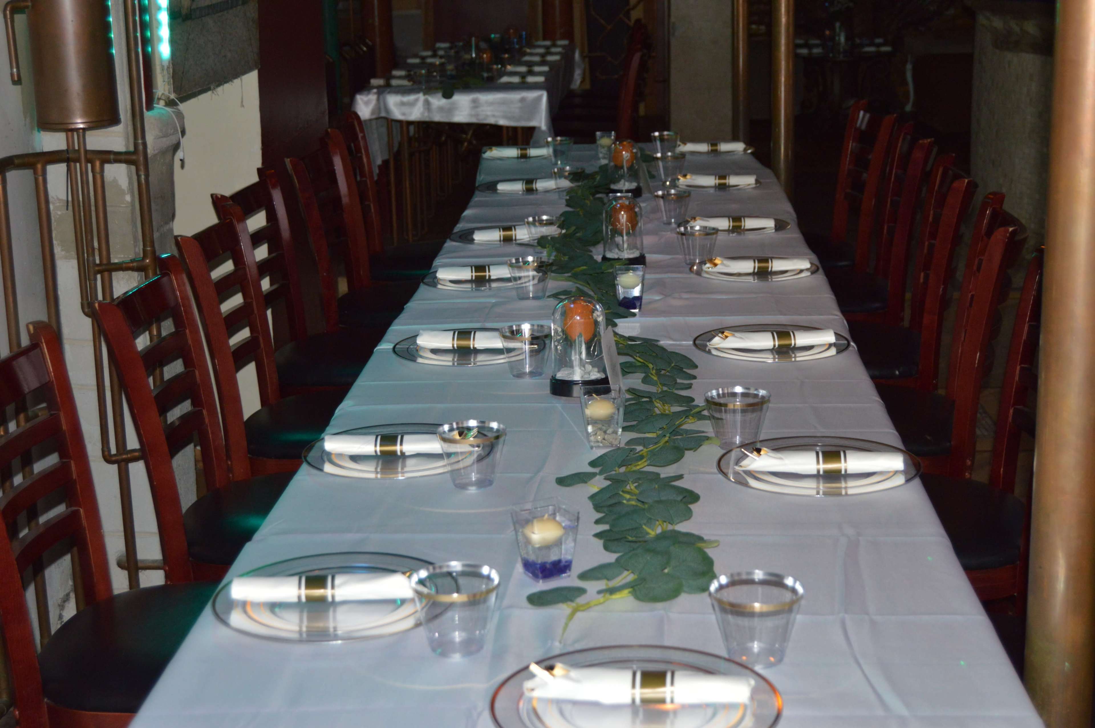 A long dining table is set with plates, glasses, and a decorative leafy center arrangement in a well-lit restaurant.