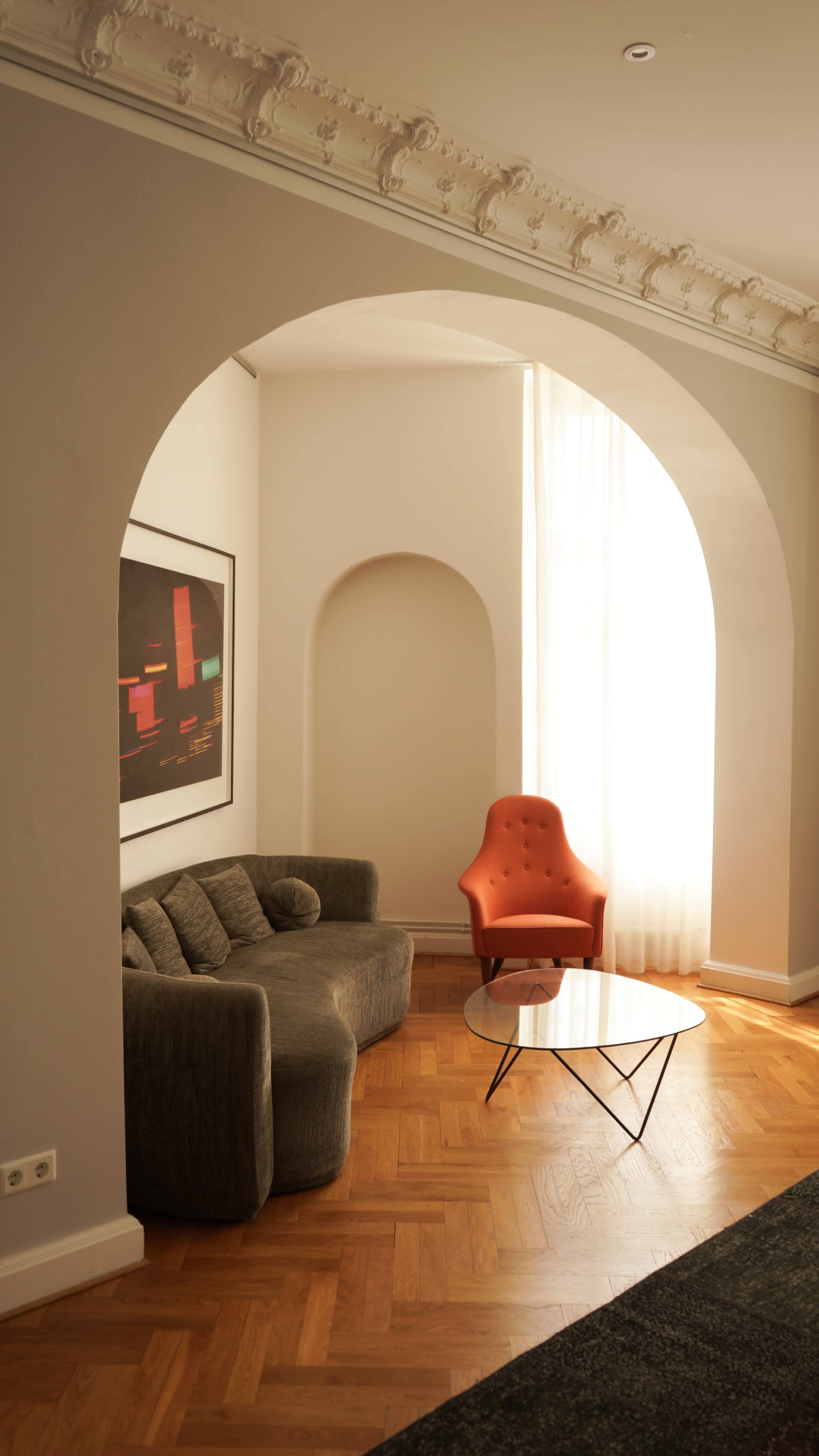 The image depicts a modern living room featuring a curved archway, a gray sectional sofa, an orange armchair, and a glass coffee table with a wooden floor.