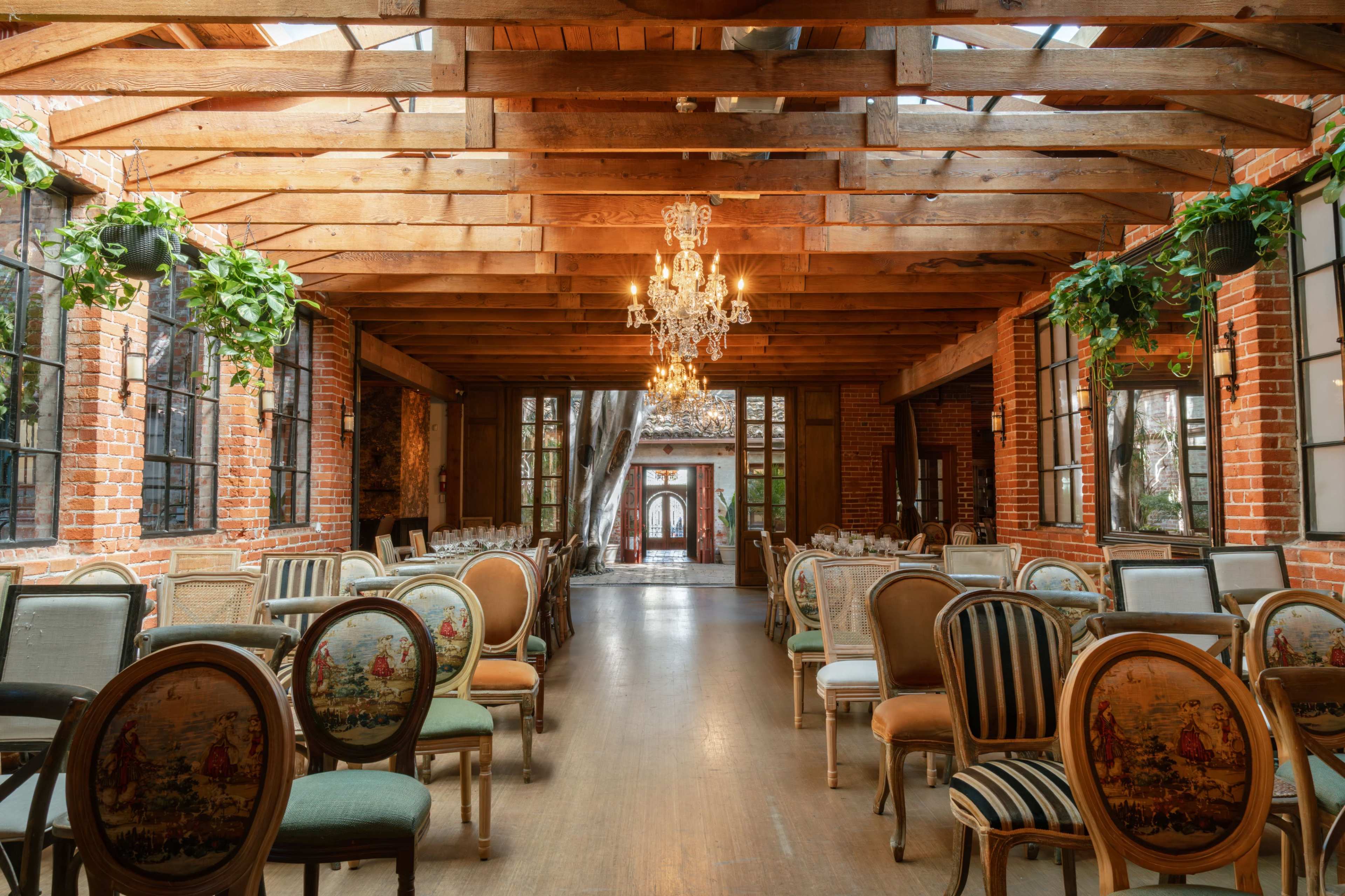 The image depicts a warm, rustic event space featuring wooden beams, brick walls, and elegant chairs arranged in rows beneath a chandelier.
