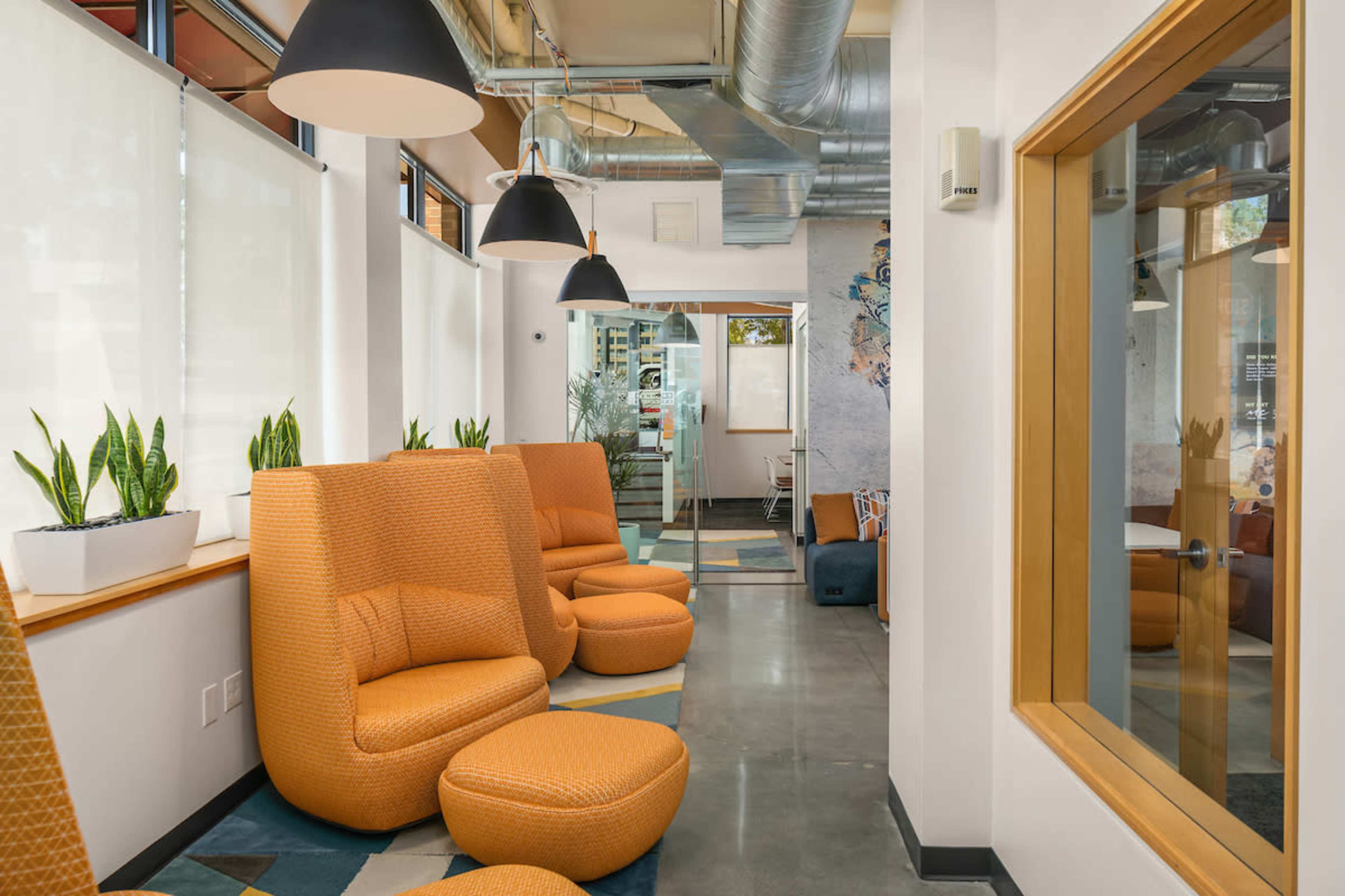 A modern, brightly lit lounge area featuring orange upholstered chairs, large pendant lights, and potted plants along the windows.
