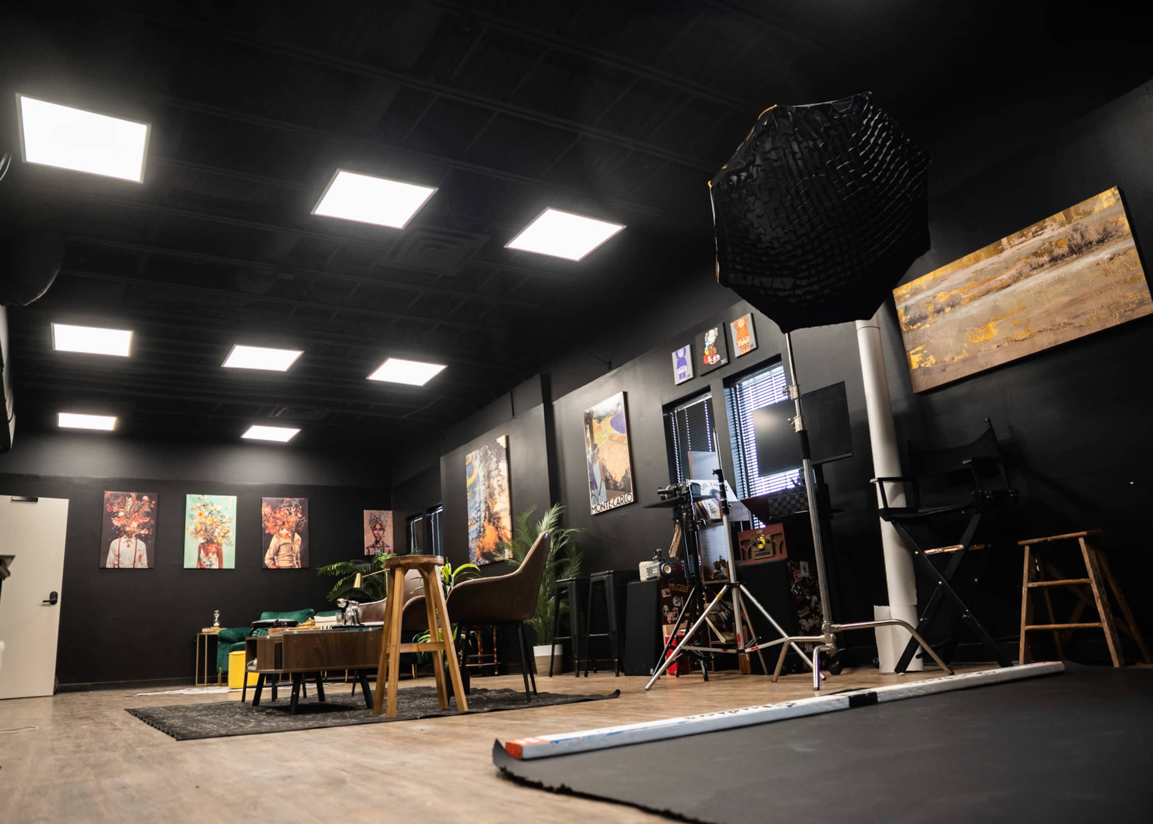The image shows a large studio space with black walls, featuring several framed artworks, a photography backdrop, and furniture arranged for a creative setting.