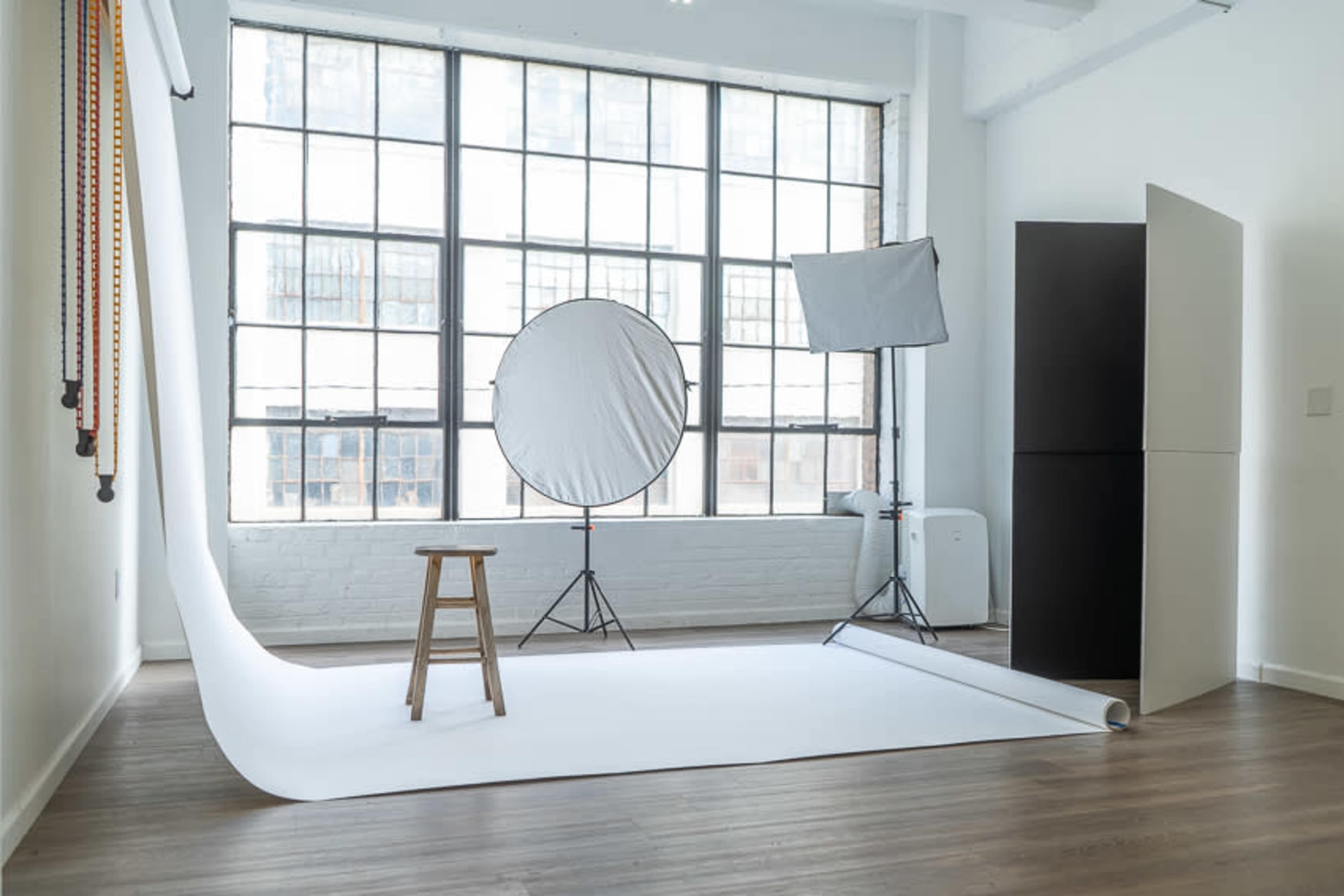 A photography studio with a white backdrop, a stool, lighting equipment, and large windows allowing natural light to enter.