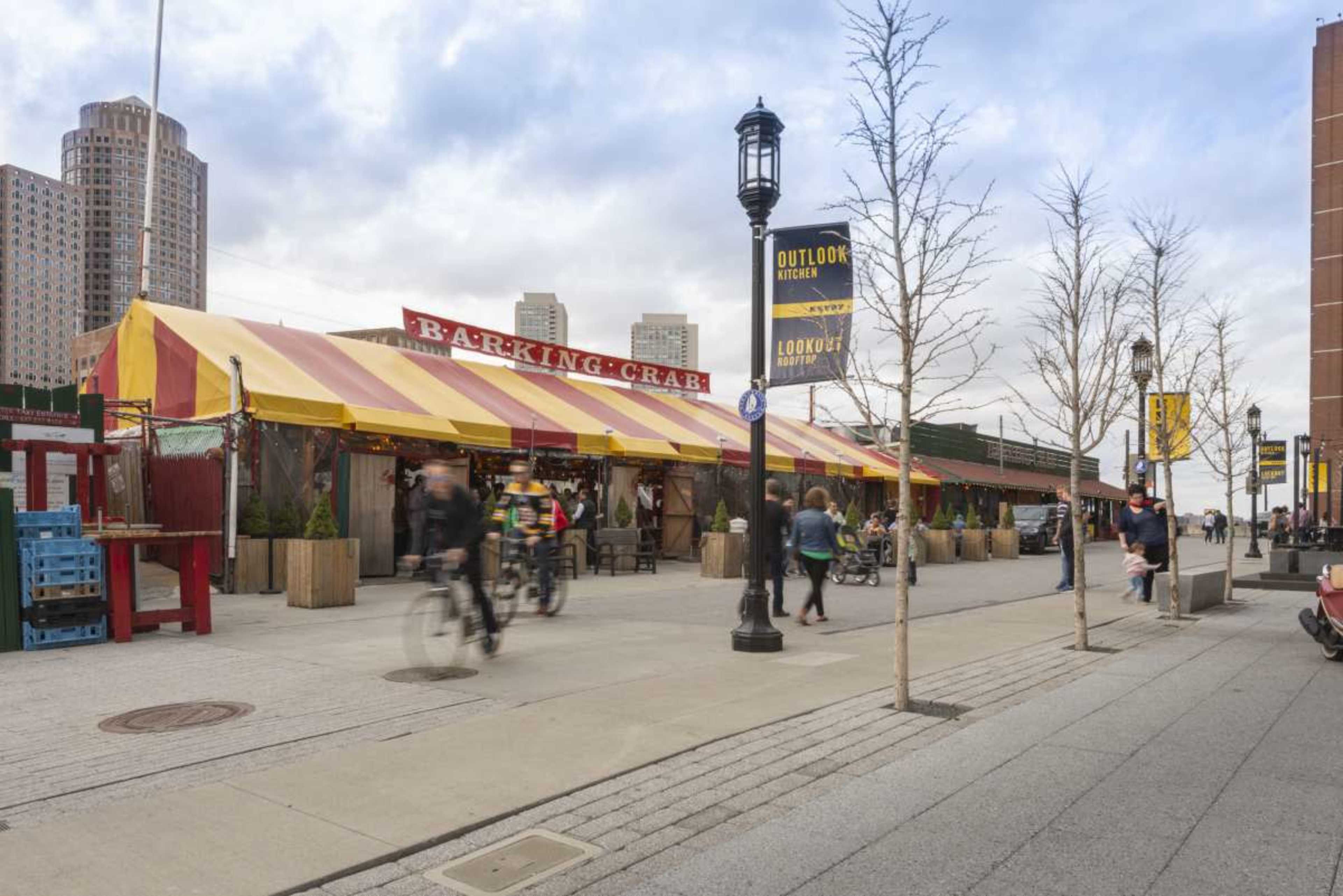 The photo shows a waterfront street scene with the colorful exterior of the Barking Crab restaurant and people walking and biking along the pathway.
