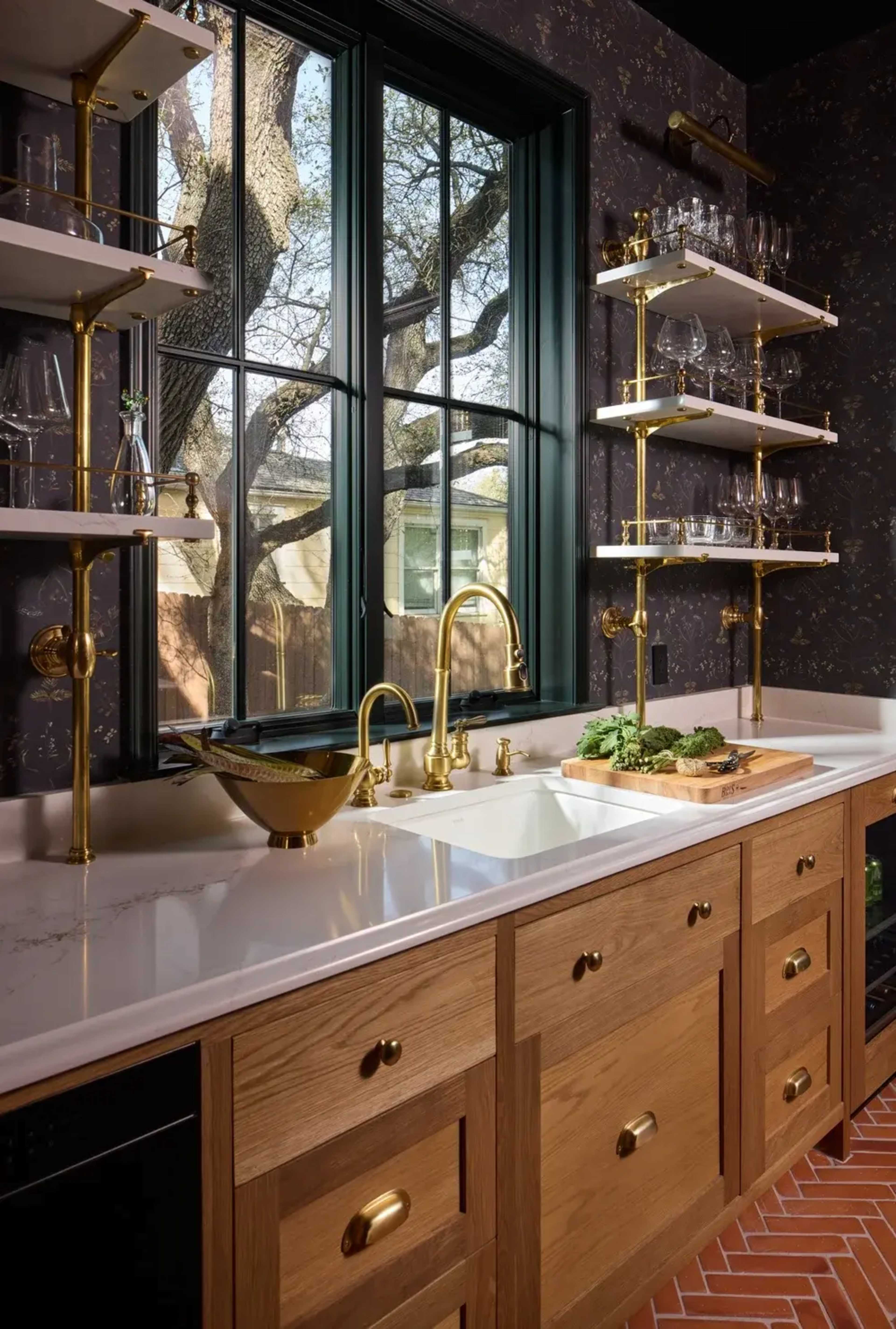 The image shows a kitchen with wooden cabinetry, a white countertop, a gold faucet, and shelves displaying glassware, illuminated by natural light through a large window.