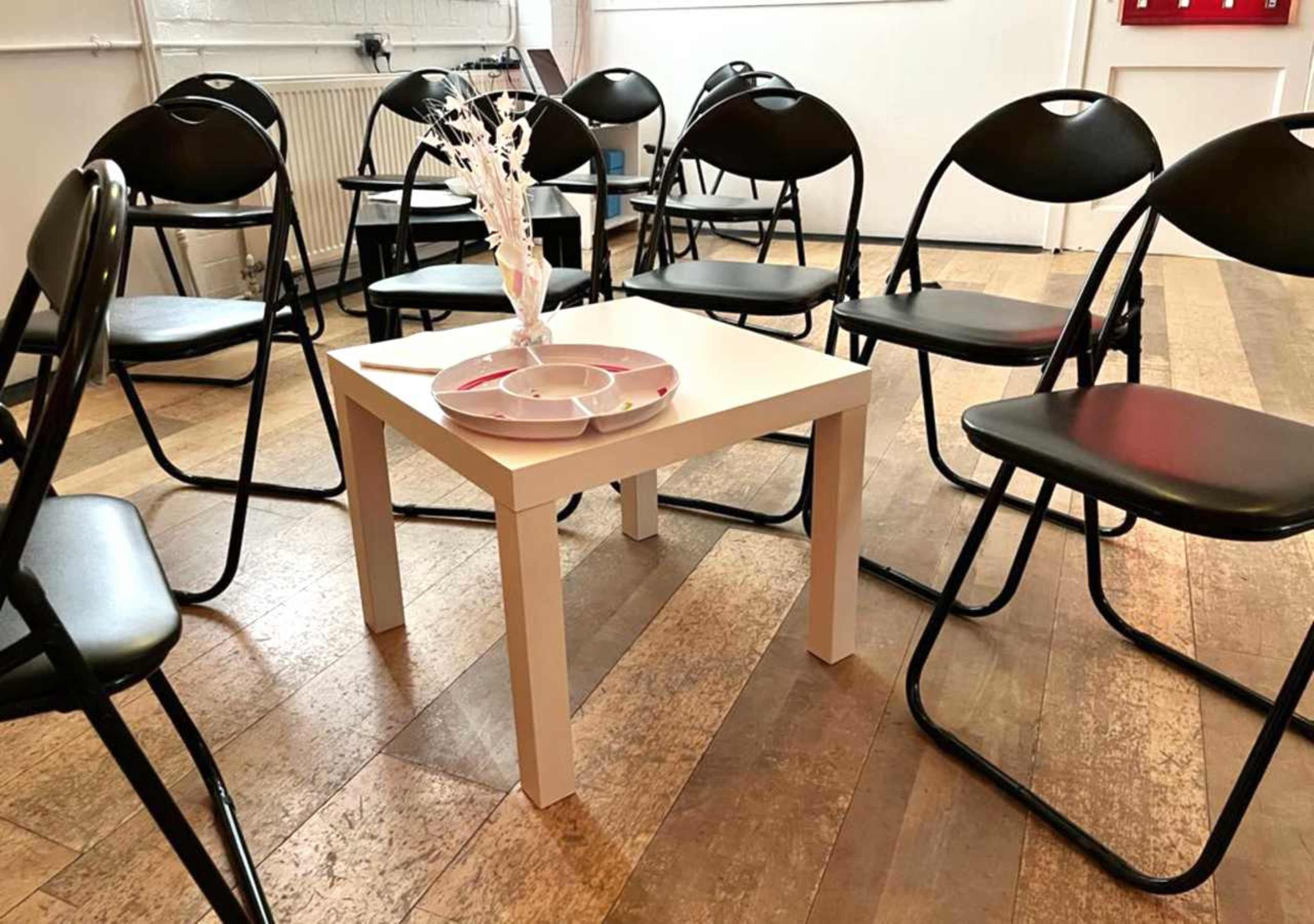A small white table is centered among several black folding chairs arranged in a circle on a wooden floor.