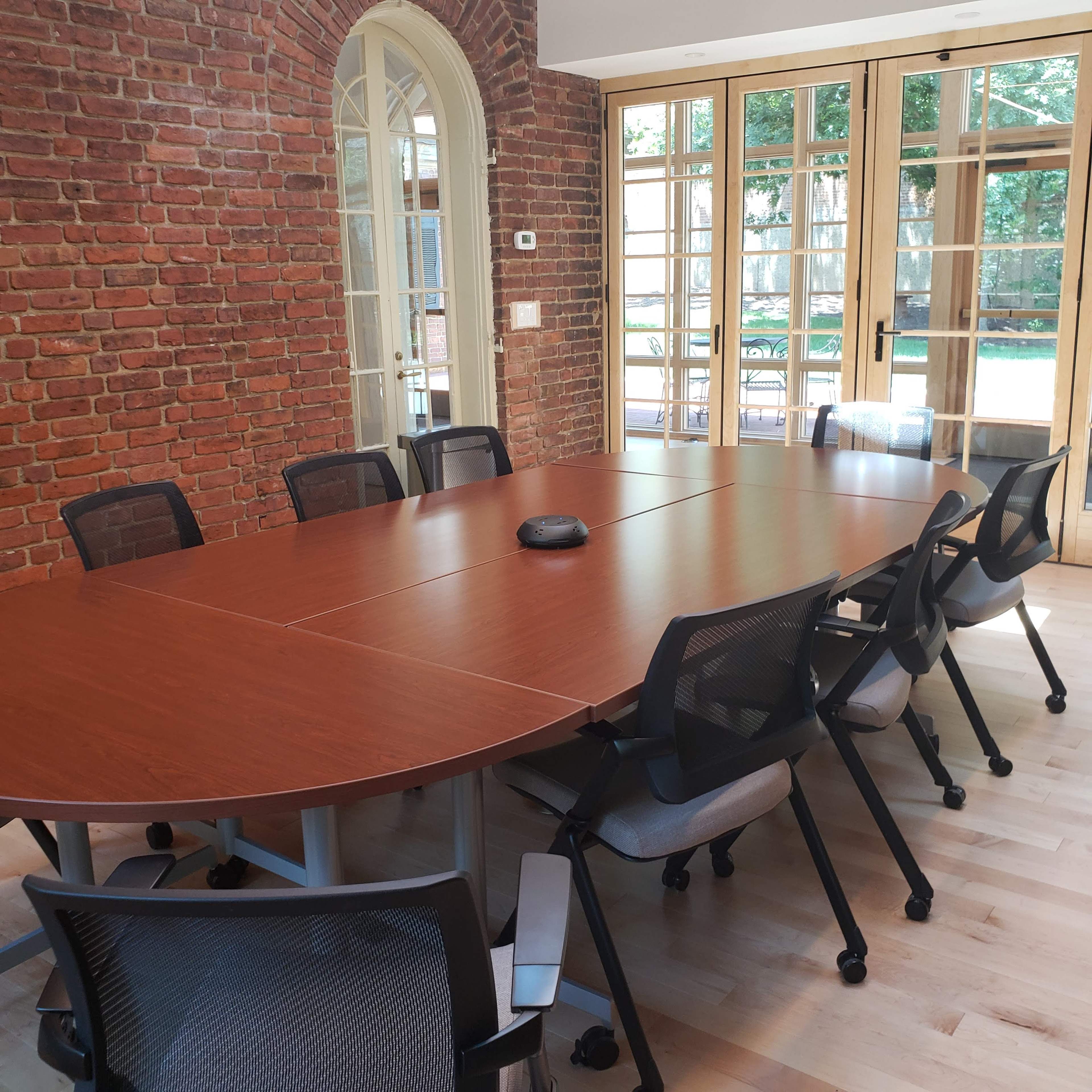 A large wooden conference table with eight rolling chairs is positioned in a room with brick walls and a view of a garden through glass doors.