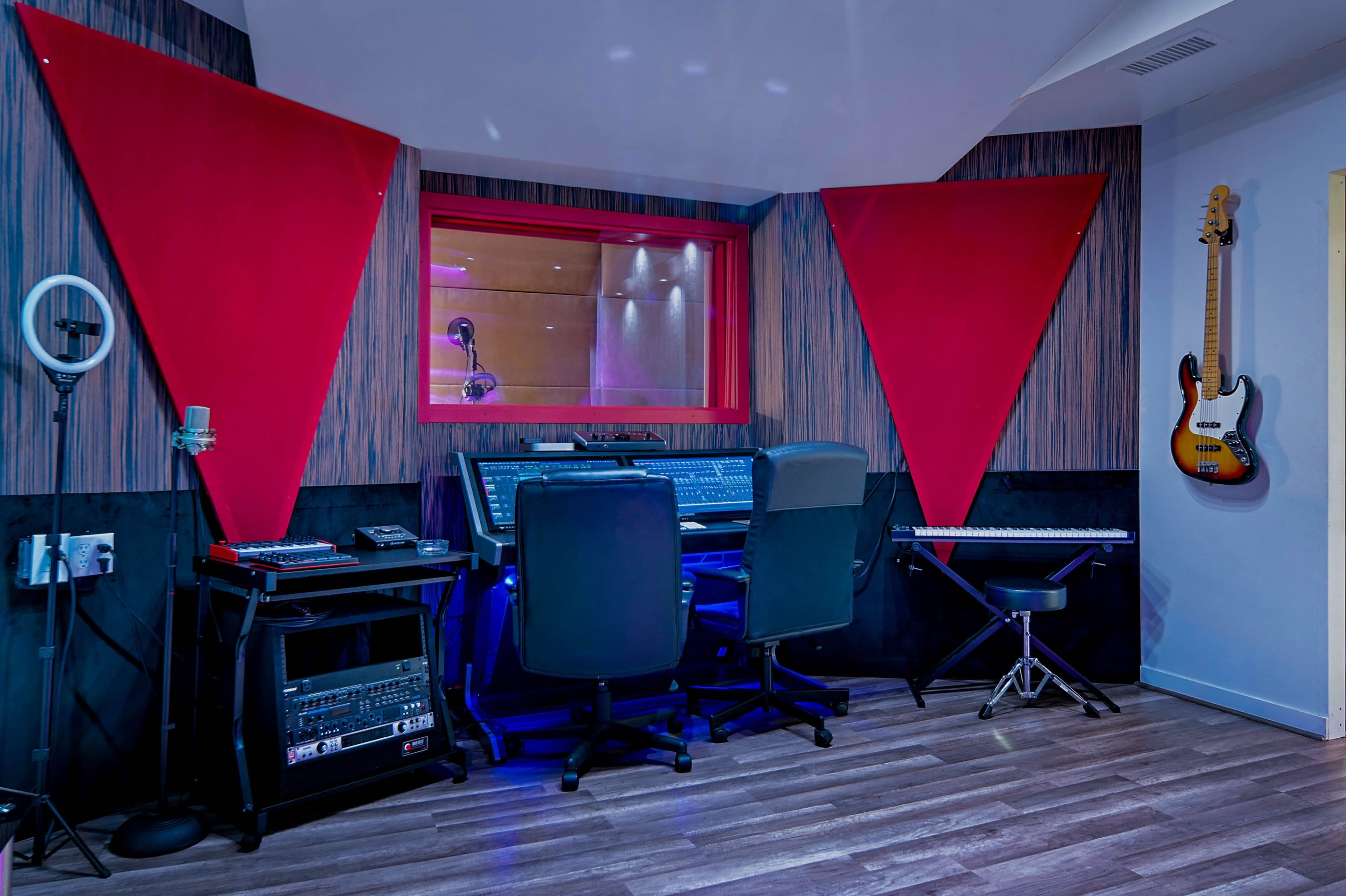 A recording studio with a control desk, microphones, and musical instruments, surrounded by soundproofing panels and a red window.