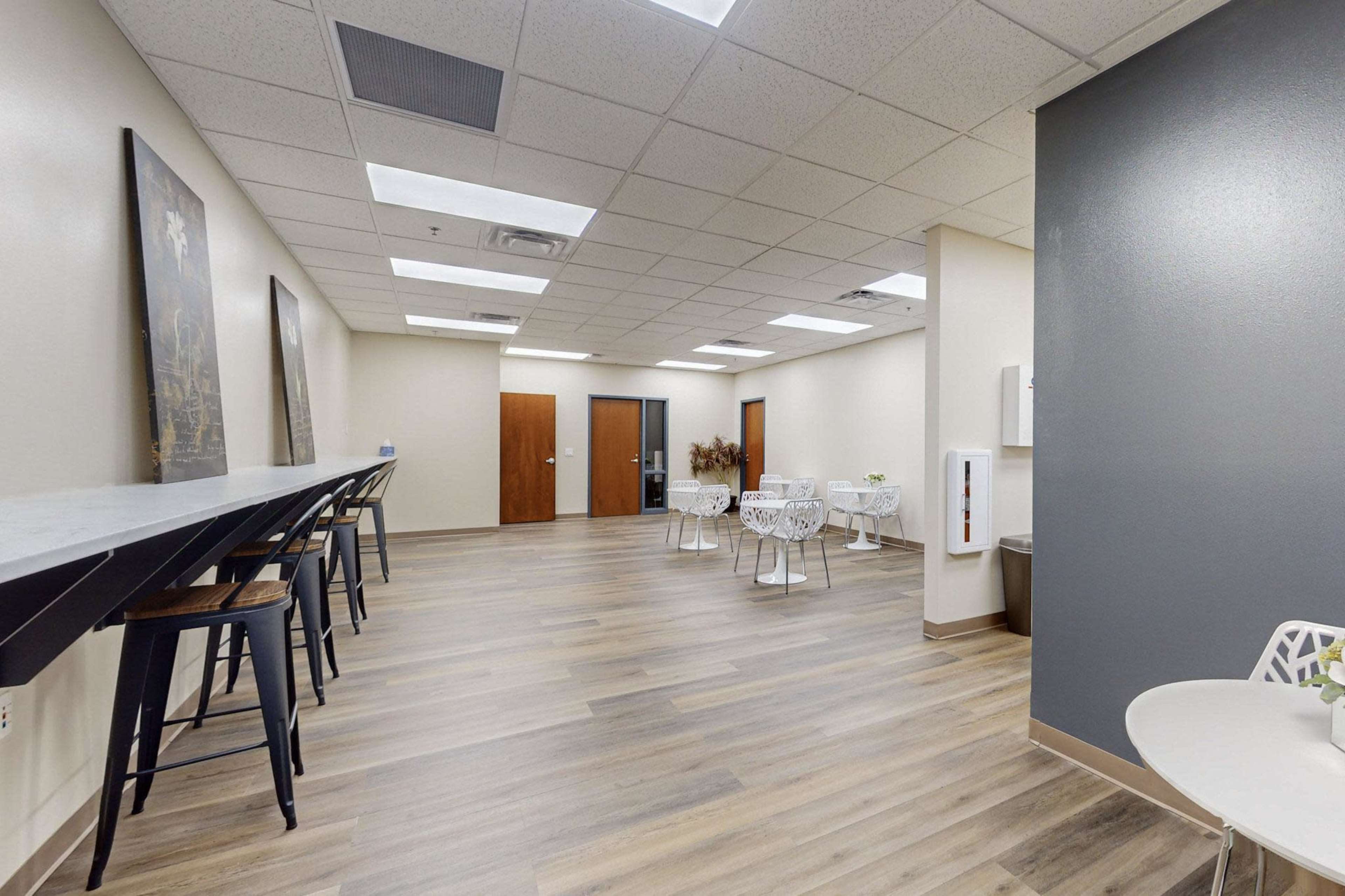 A modern waiting area with a long bar-style table, several small tables and chairs, and neutral-colored walls.